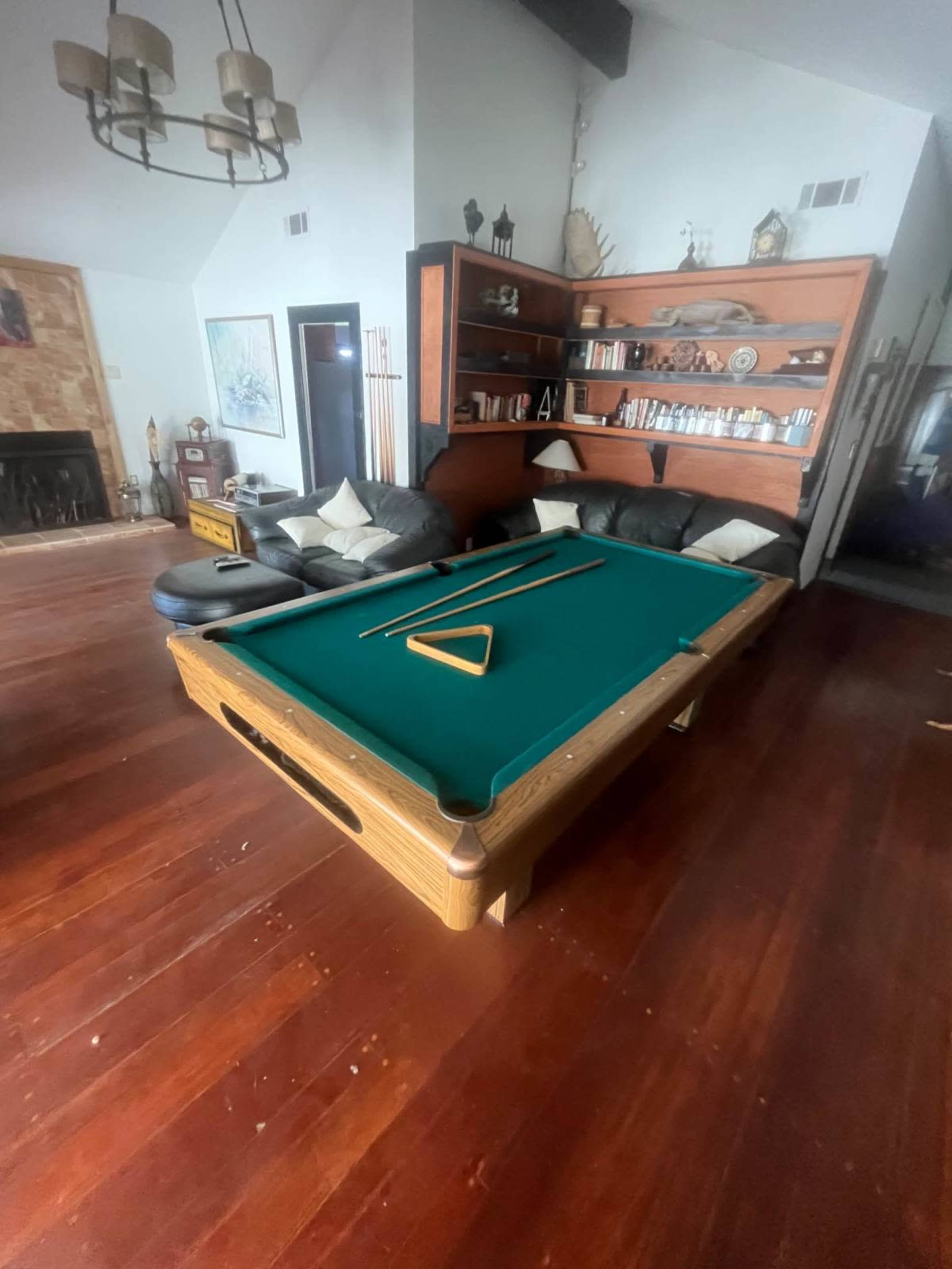 The image shows a game room featuring a pool table with a green felt surface, surrounded by dark leather couches and wooden shelves filled with books and decorative items.