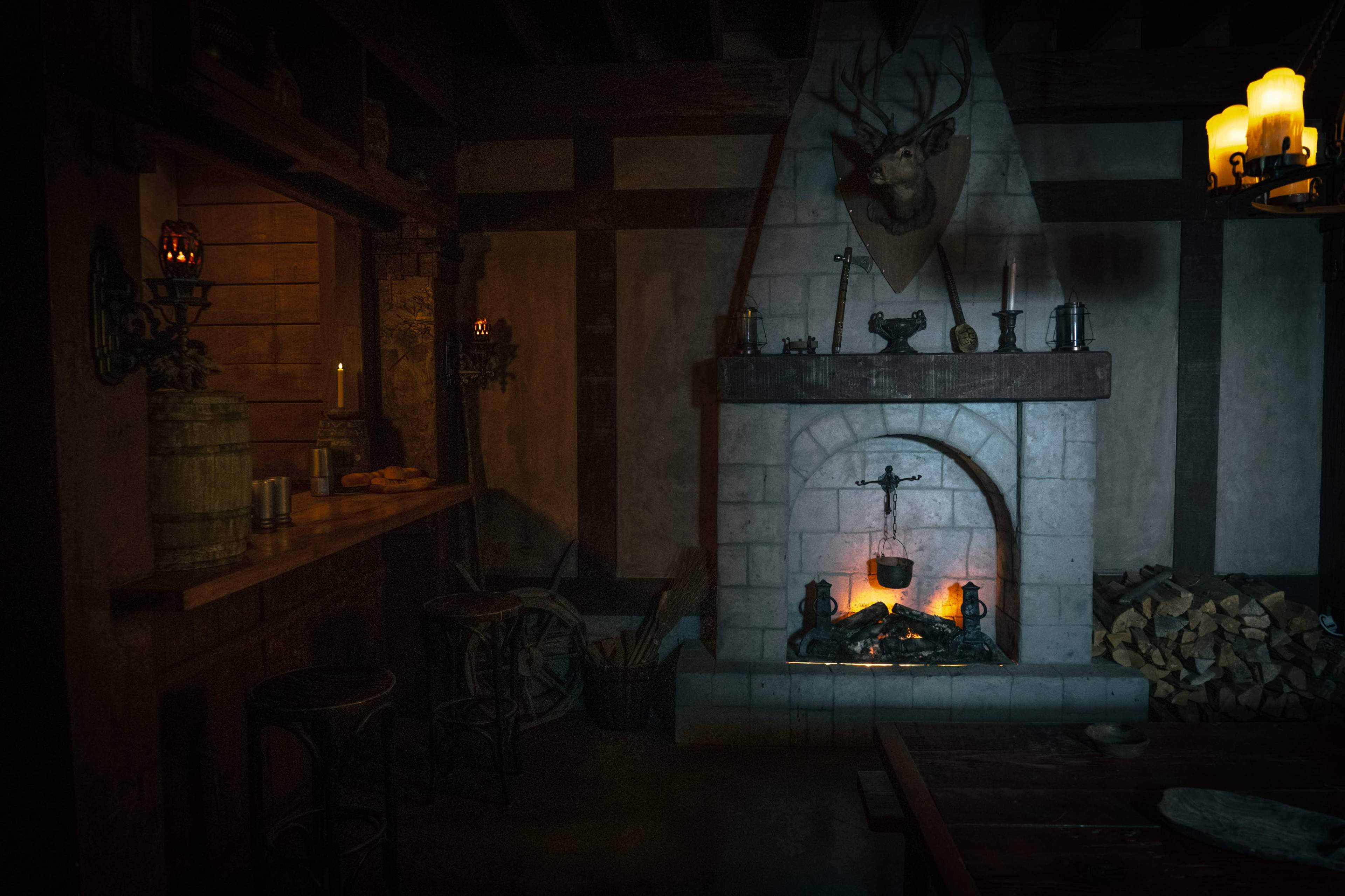 The image shows a dimly lit rustic interior featuring a stone fireplace with a roaring fire, wooden beams, and a bar area with stools.