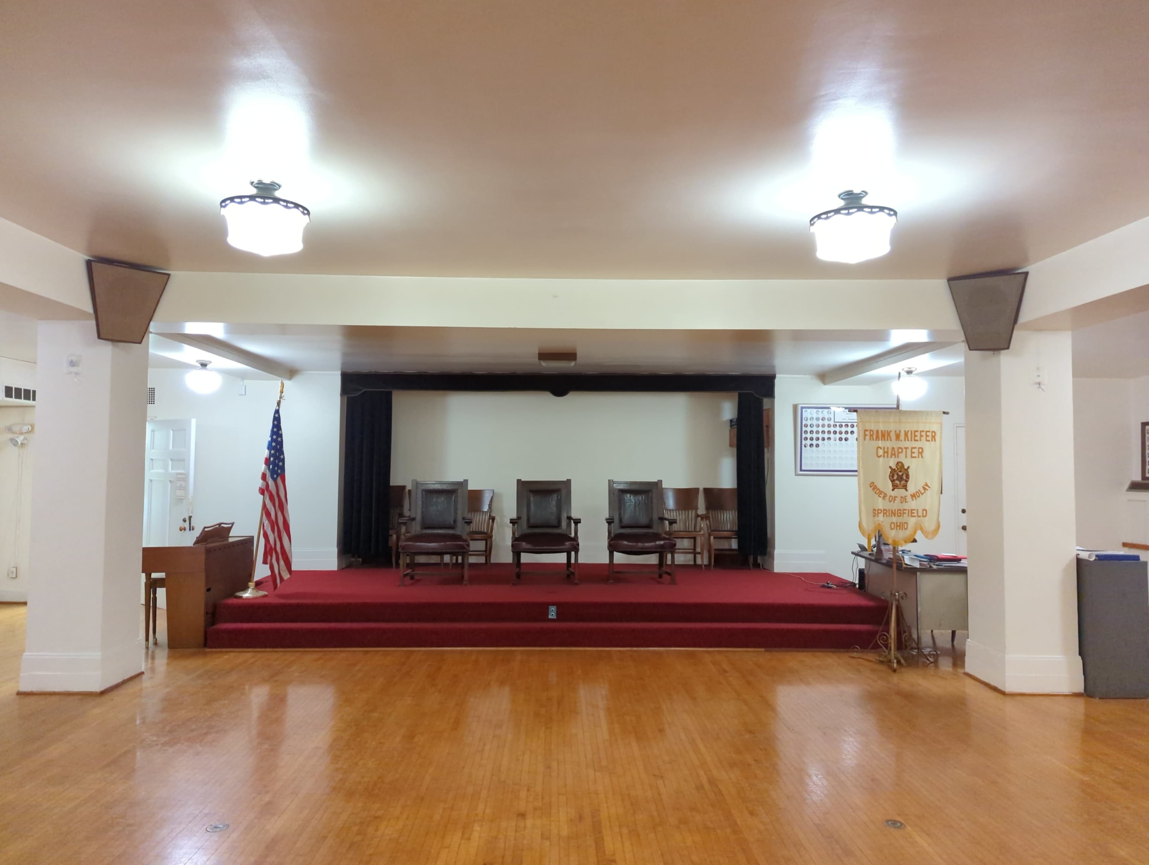 A spacious hall with a red carpet, three wooden chairs on an elevated platform, an American flag on the left, and a side table with a sign and various items on the right.