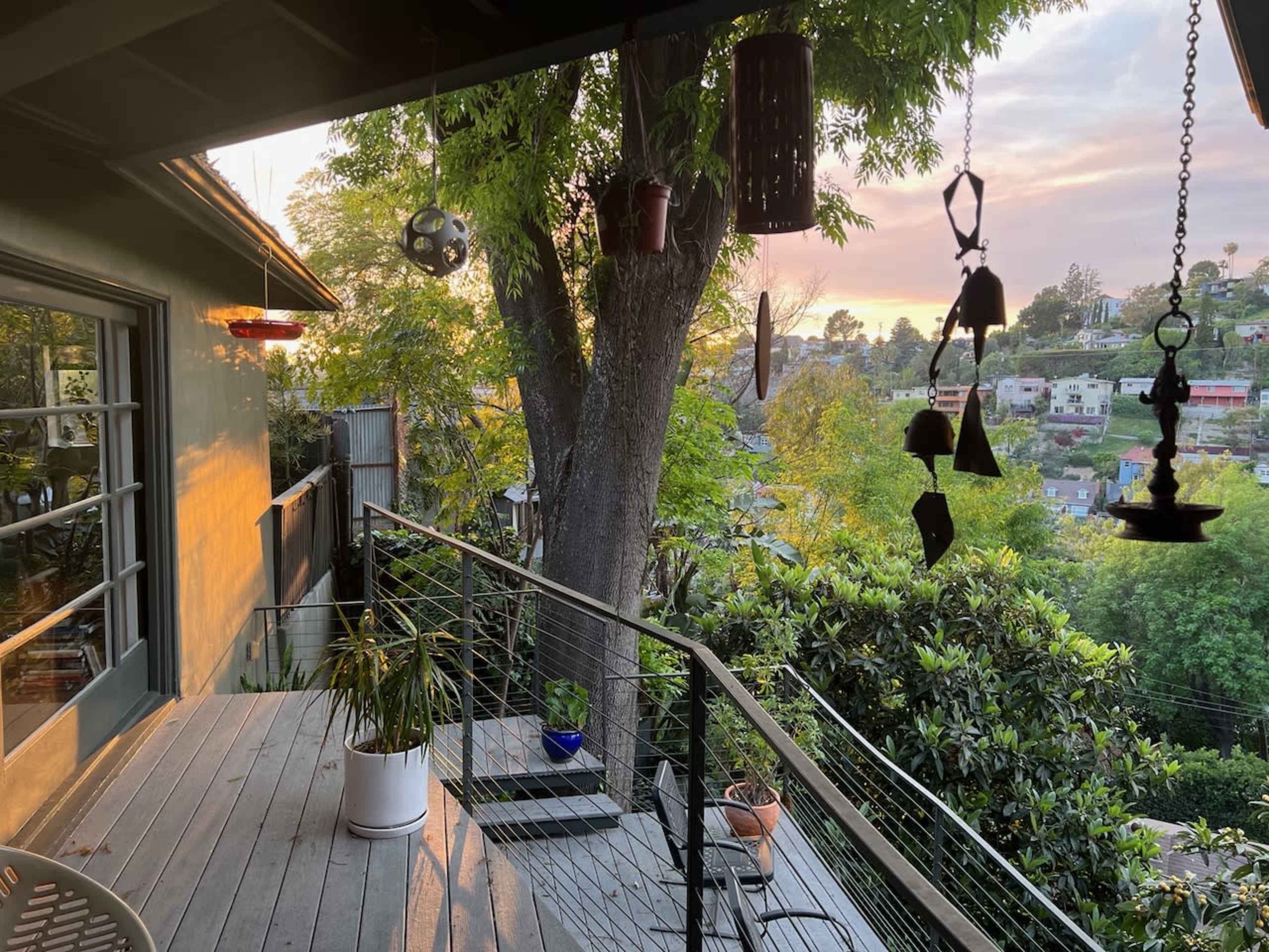 A wooden deck is surrounded by hanging decorations and overlooks a neighborhood with sunset lighting filtering through the trees.