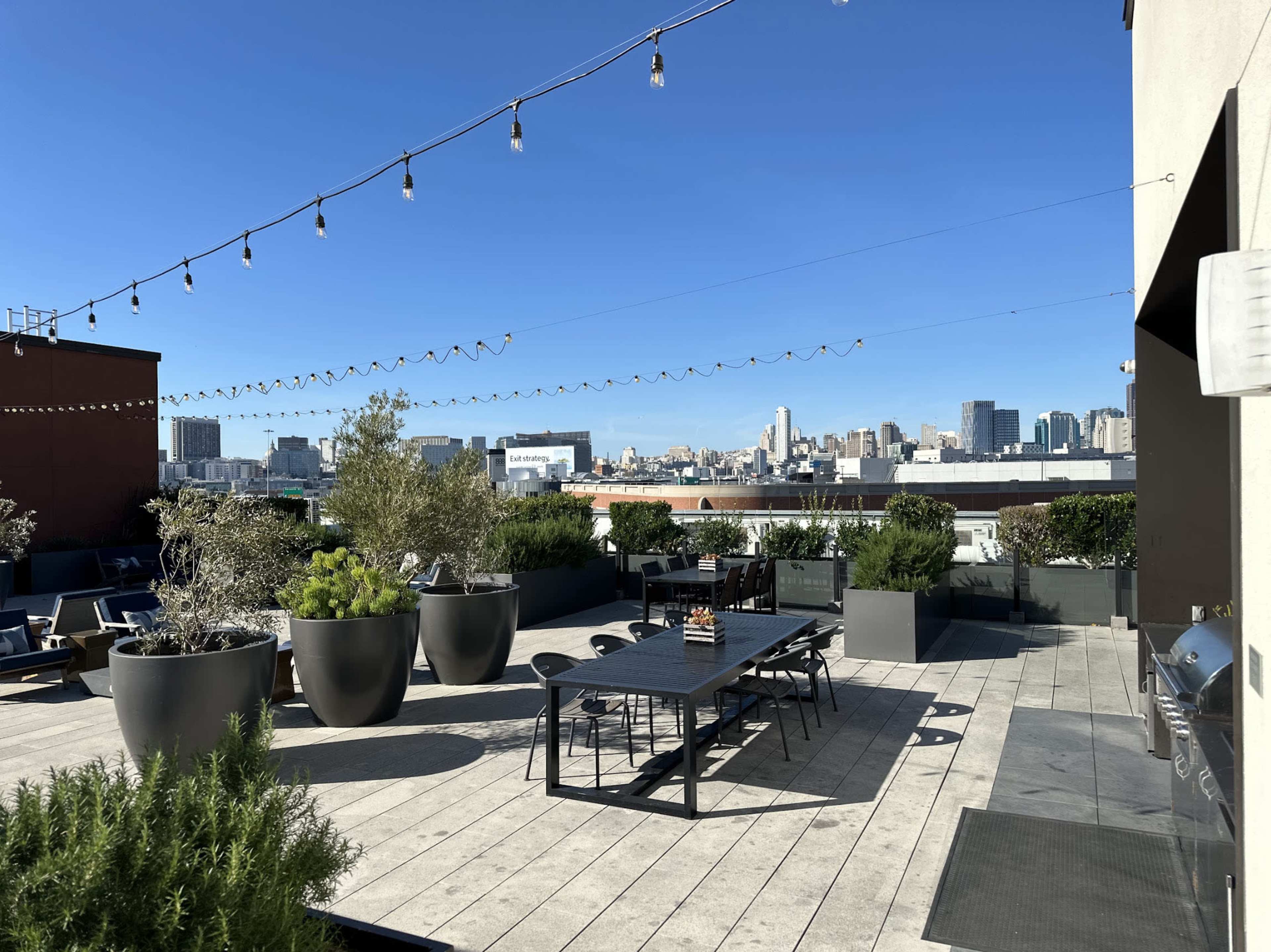 A rooftop terrace features a large dining table surrounded by potted plants and lounge chairs, with a city skyline visible in the background under a clear blue sky.