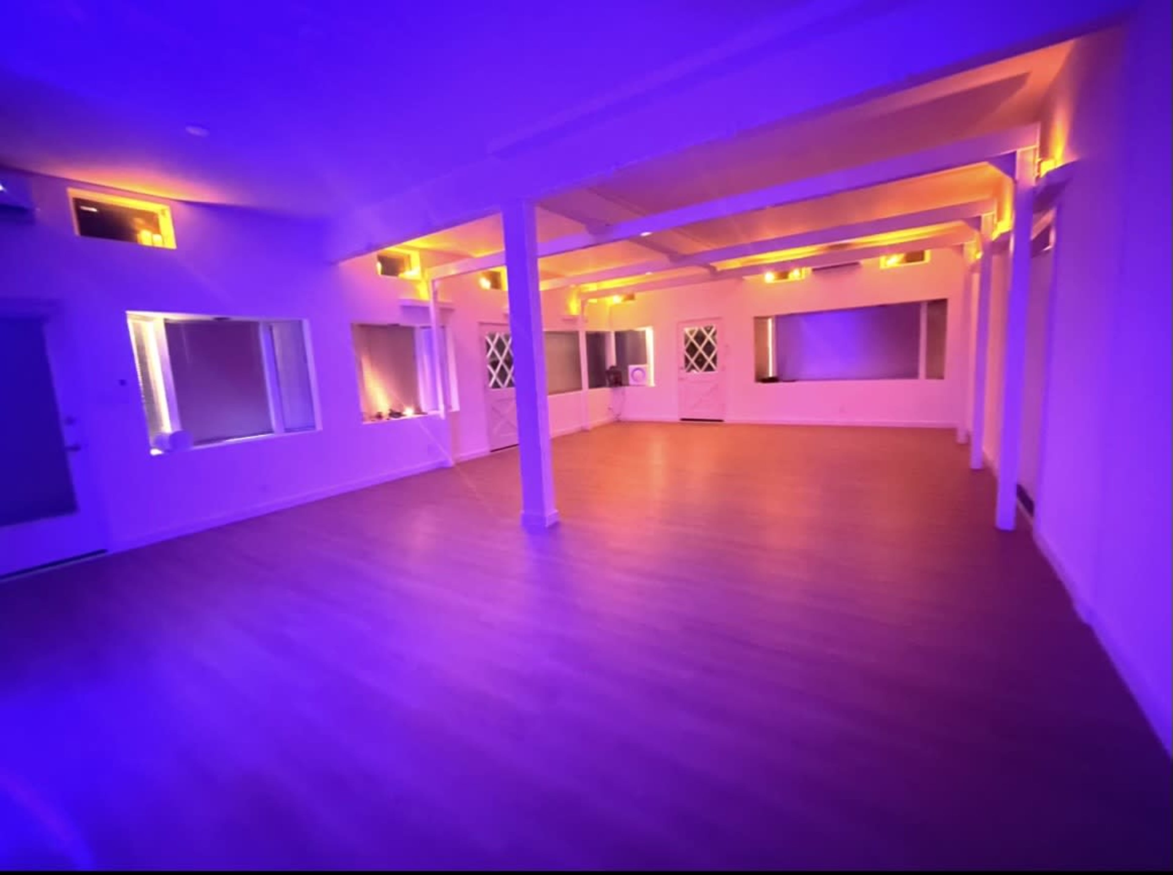 The image shows a spacious room illuminated by purple and orange lighting, with wooden floors and large windows.
