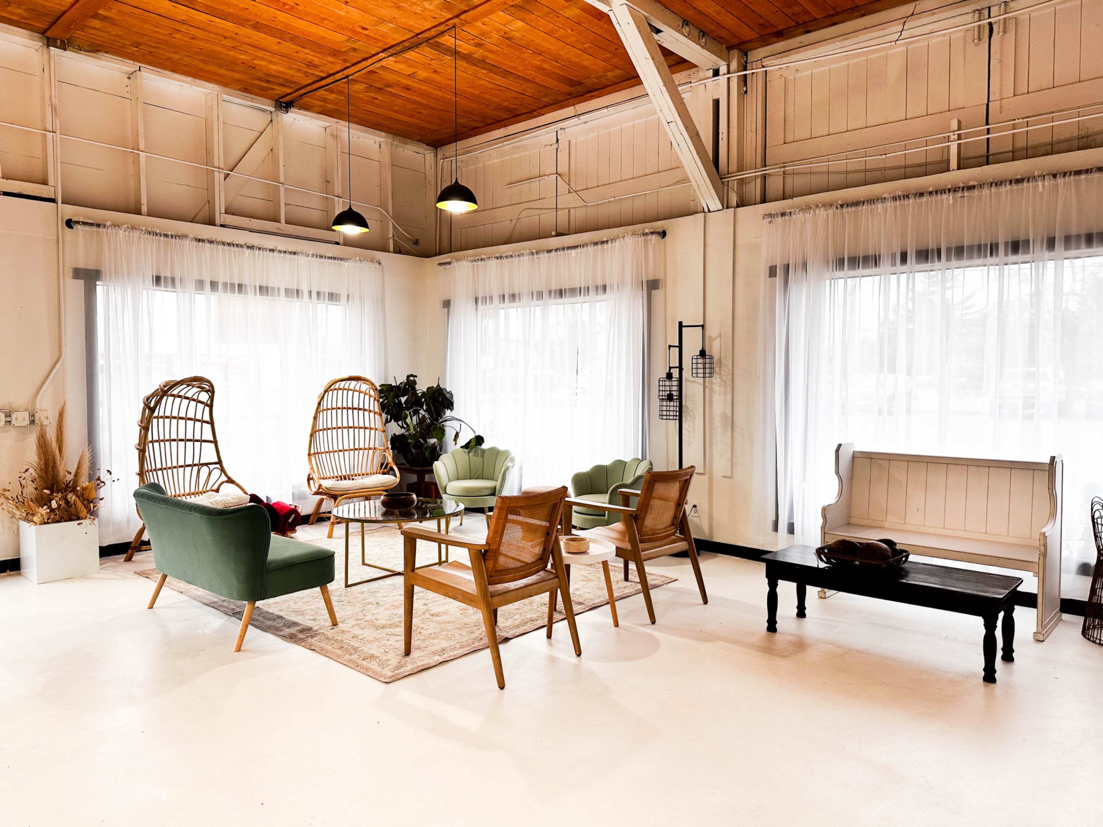 The image shows a spacious interior with a mix of seating options, including rattan chairs and modern green armchairs, surrounded by large windows with sheer curtains.