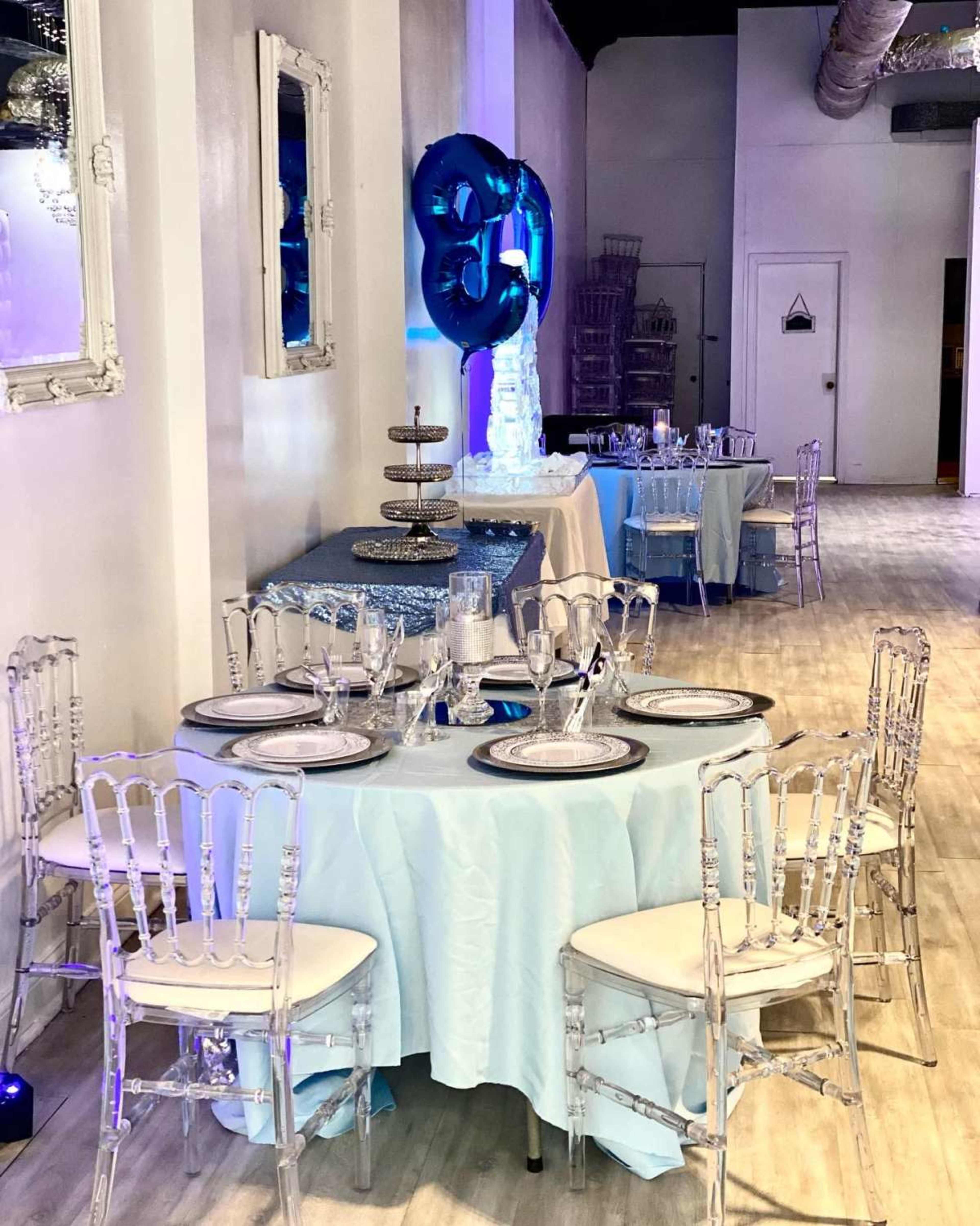 The image shows a decorated dining area with tables set with light blue tablecloths, clear chiavari chairs, and elegant tableware, complemented by a balloon display in the background.