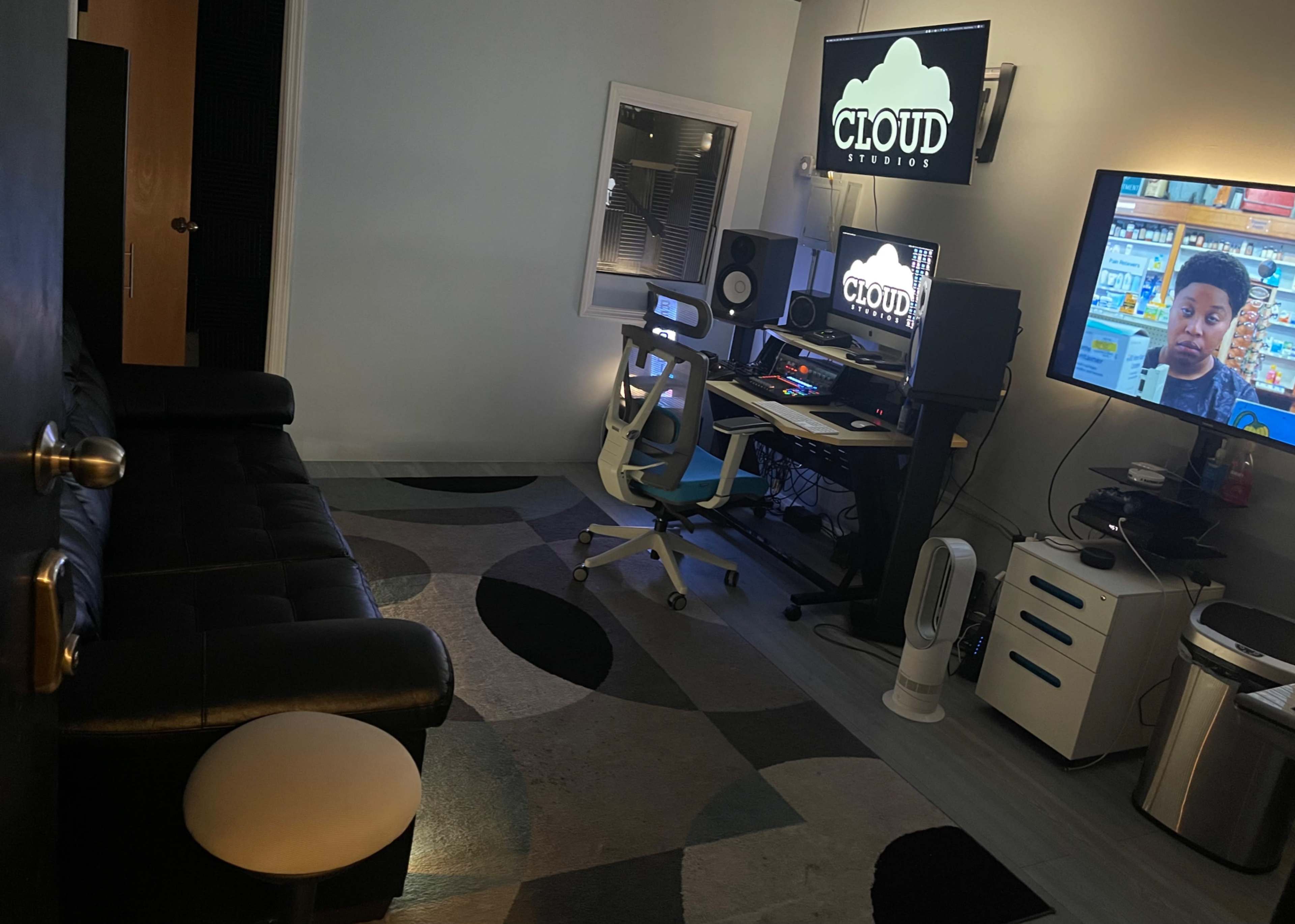 A small studio space with a computer workstation, a television, a black couch, and various electronic equipment arranged on a patterned rug.