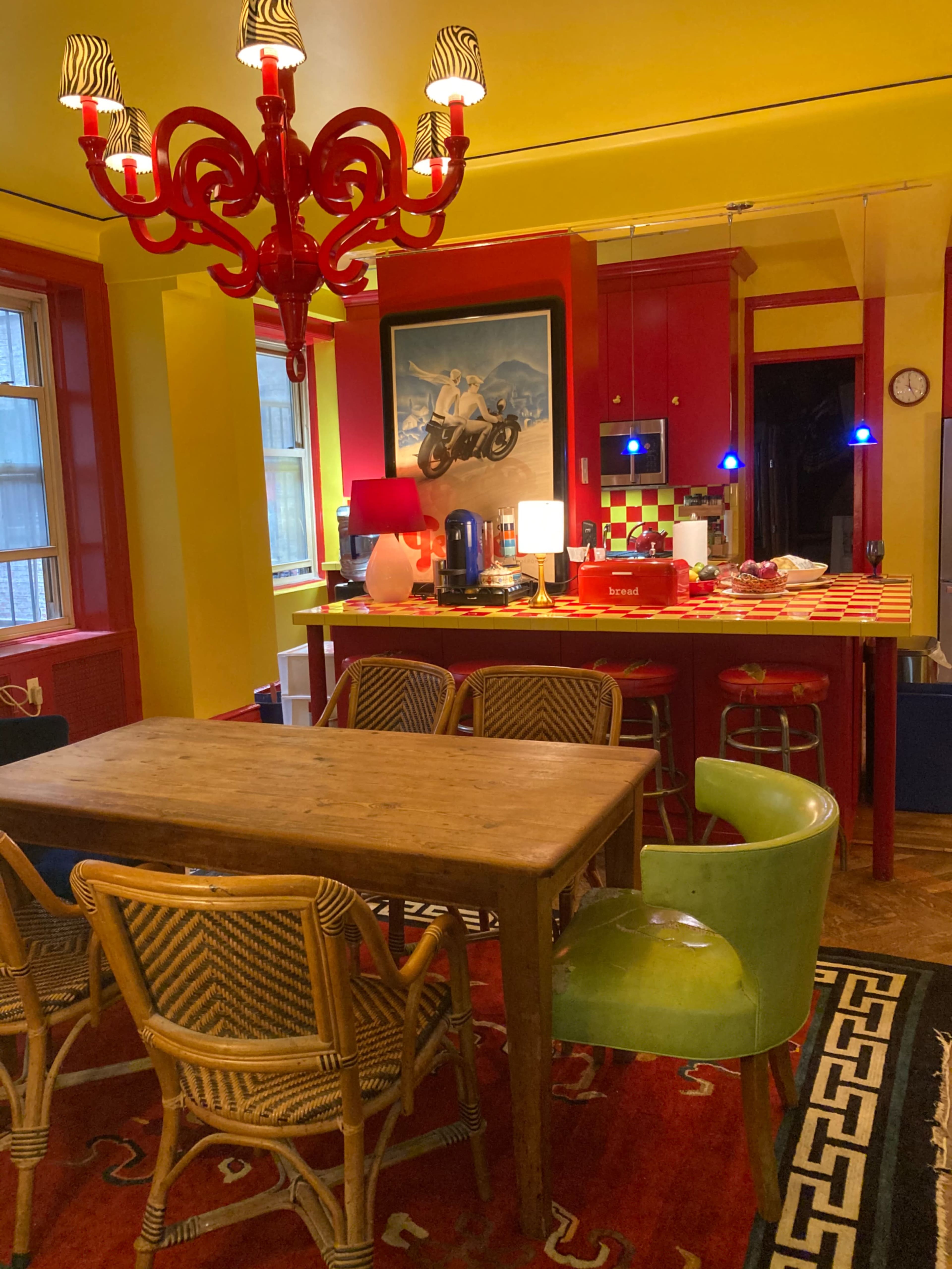 A brightly colored kitchen and dining area with a wooden table surrounded by rattan chairs, a red chandelier, and vibrant yellow walls.