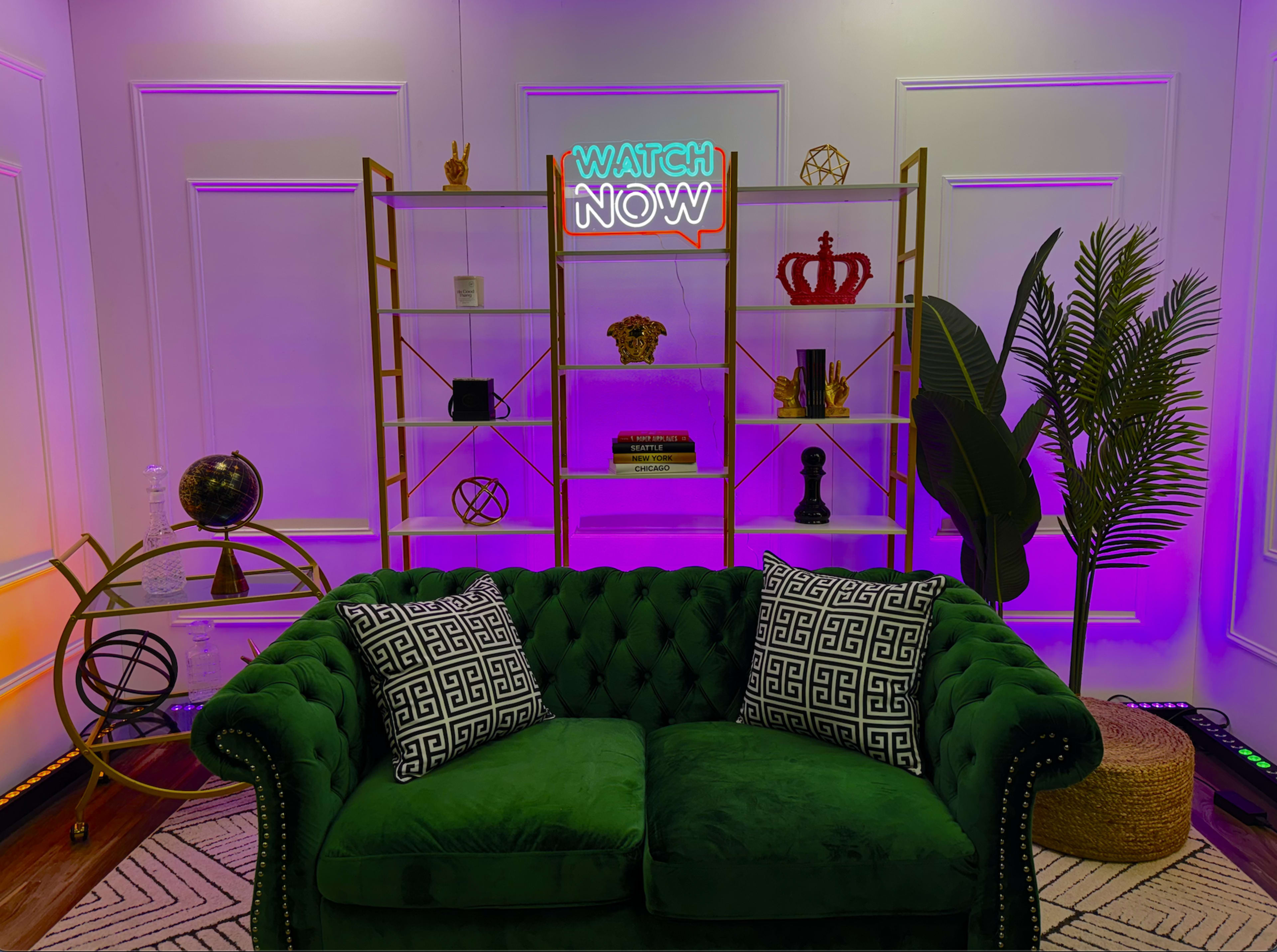 A green velvet couch with patterned pillows in front of a decorative bookshelf and a neon sign that says "WATCH NOW."
