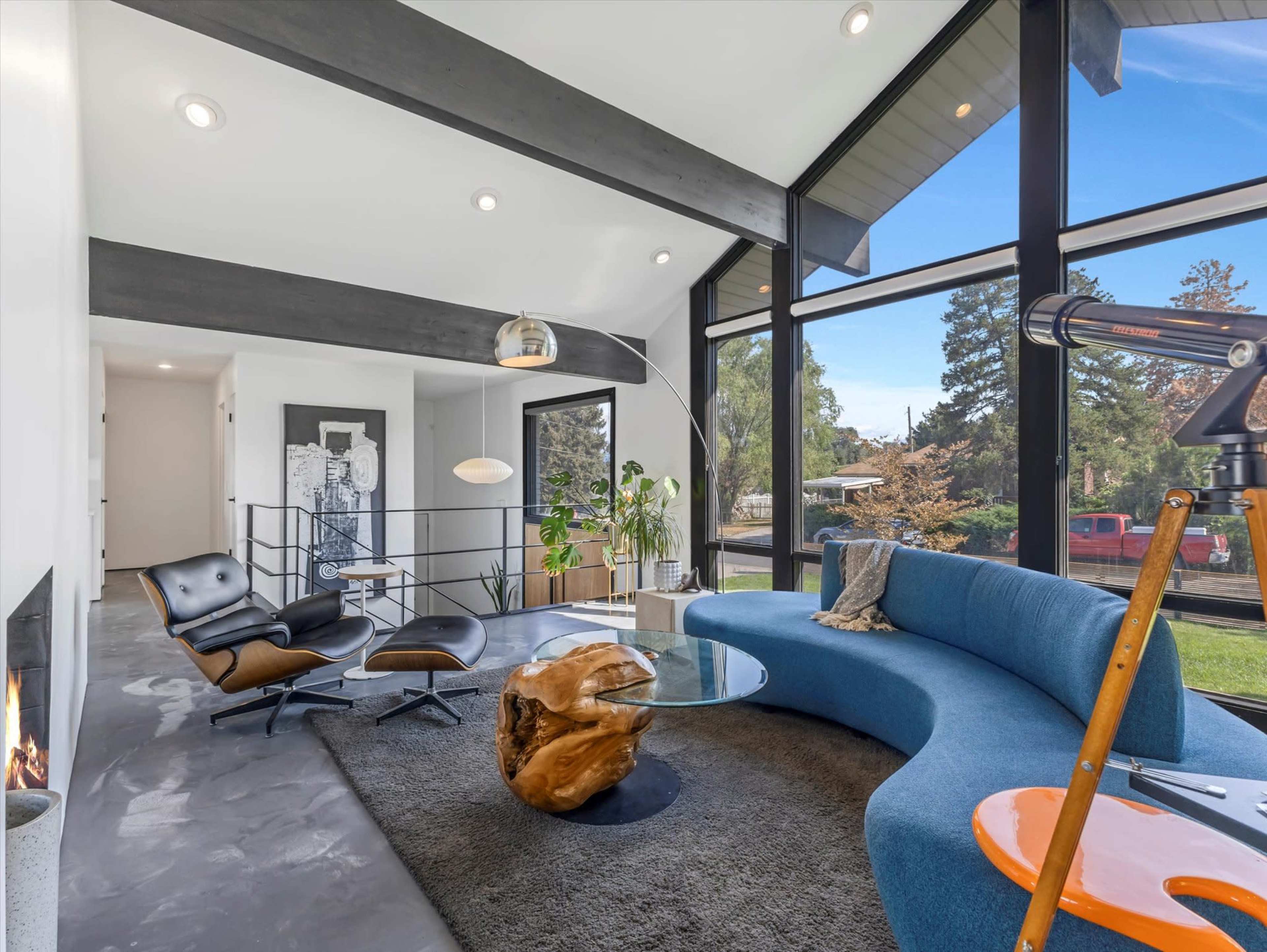The image shows a modern living room with a curved blue sofa, a round glass coffee table, a black lounge chair, and large windows allowing natural light and showcasing the outdoor scenery.