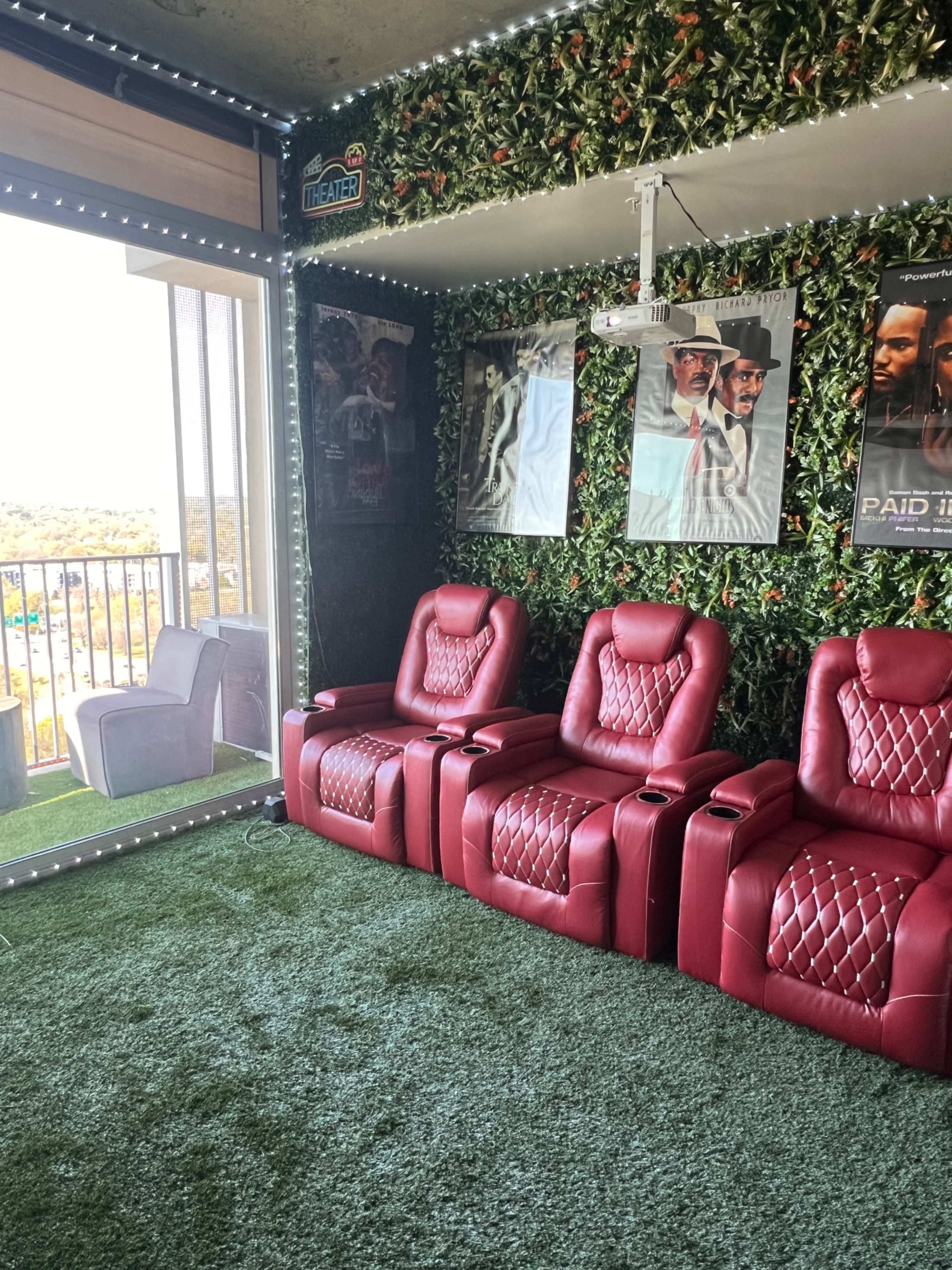 The image shows a home theater with red reclining chairs, a projector, and a wall adorned with movie posters and greenery.