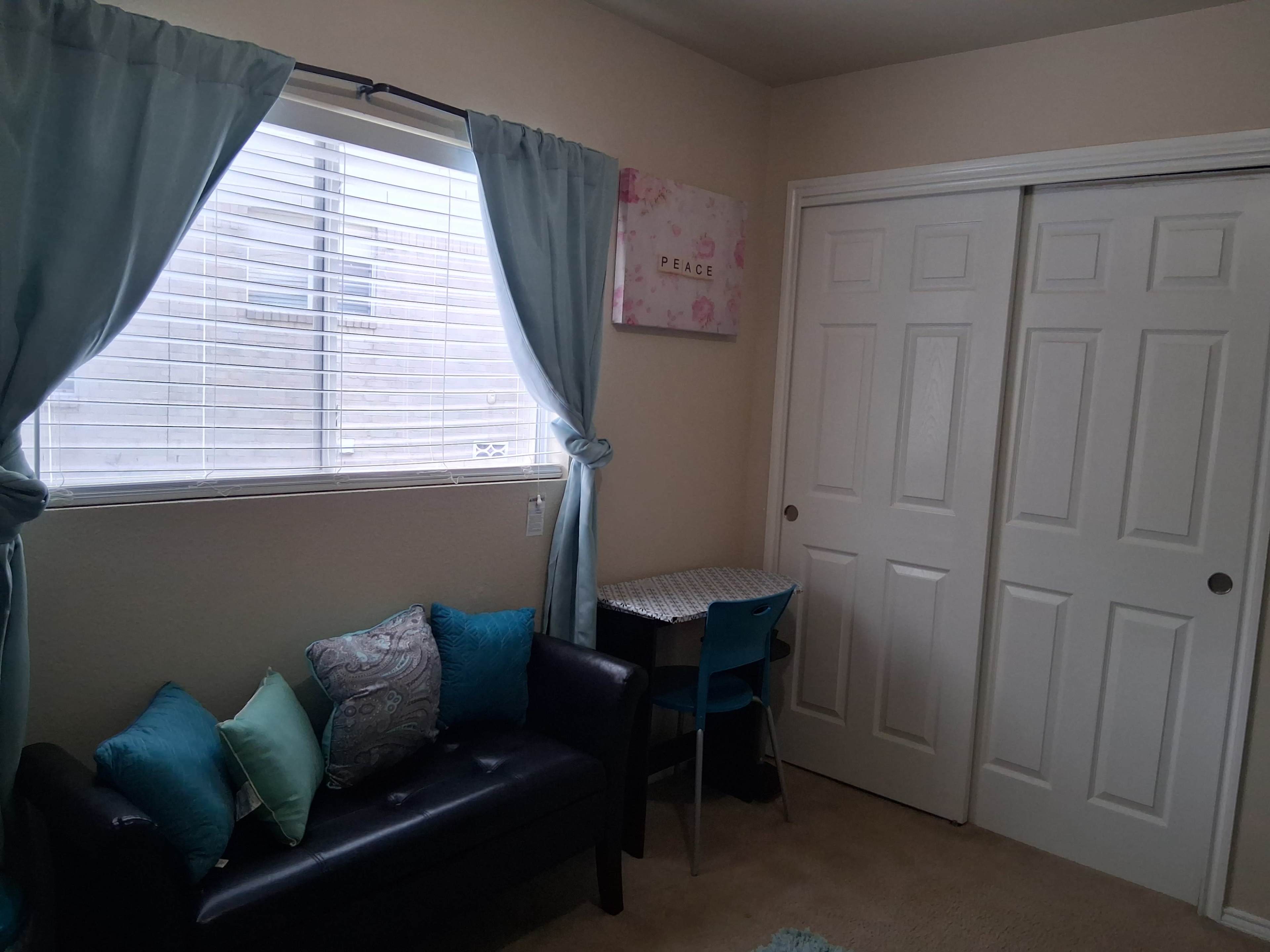 The image shows a small room with a black couch, a blue chair and desk, a window with light blue curtains, and two white closet doors.