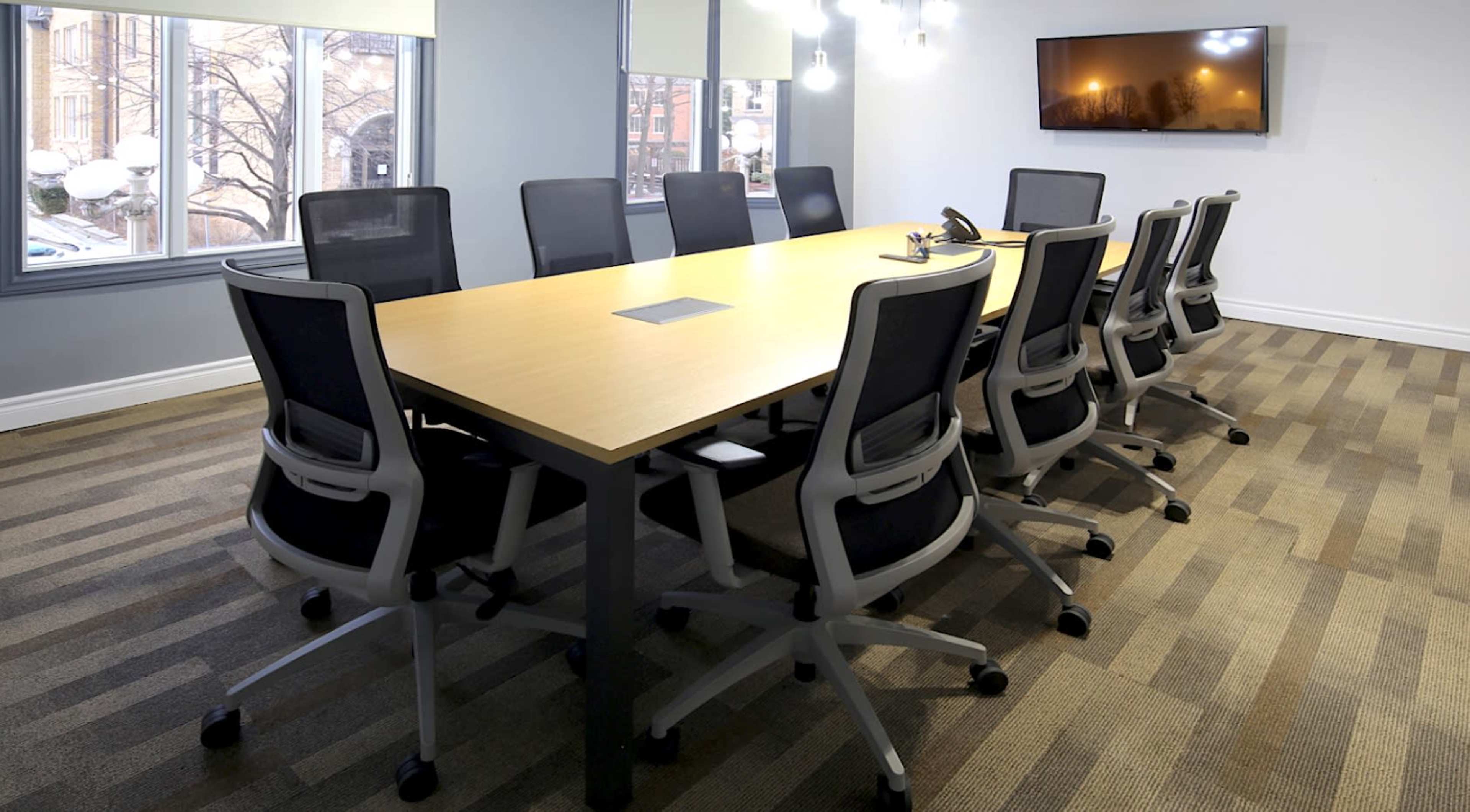 A modern conference room features a long wooden table surrounded by black ergonomic chairs, with large windows and a flat-screen television on the wall.