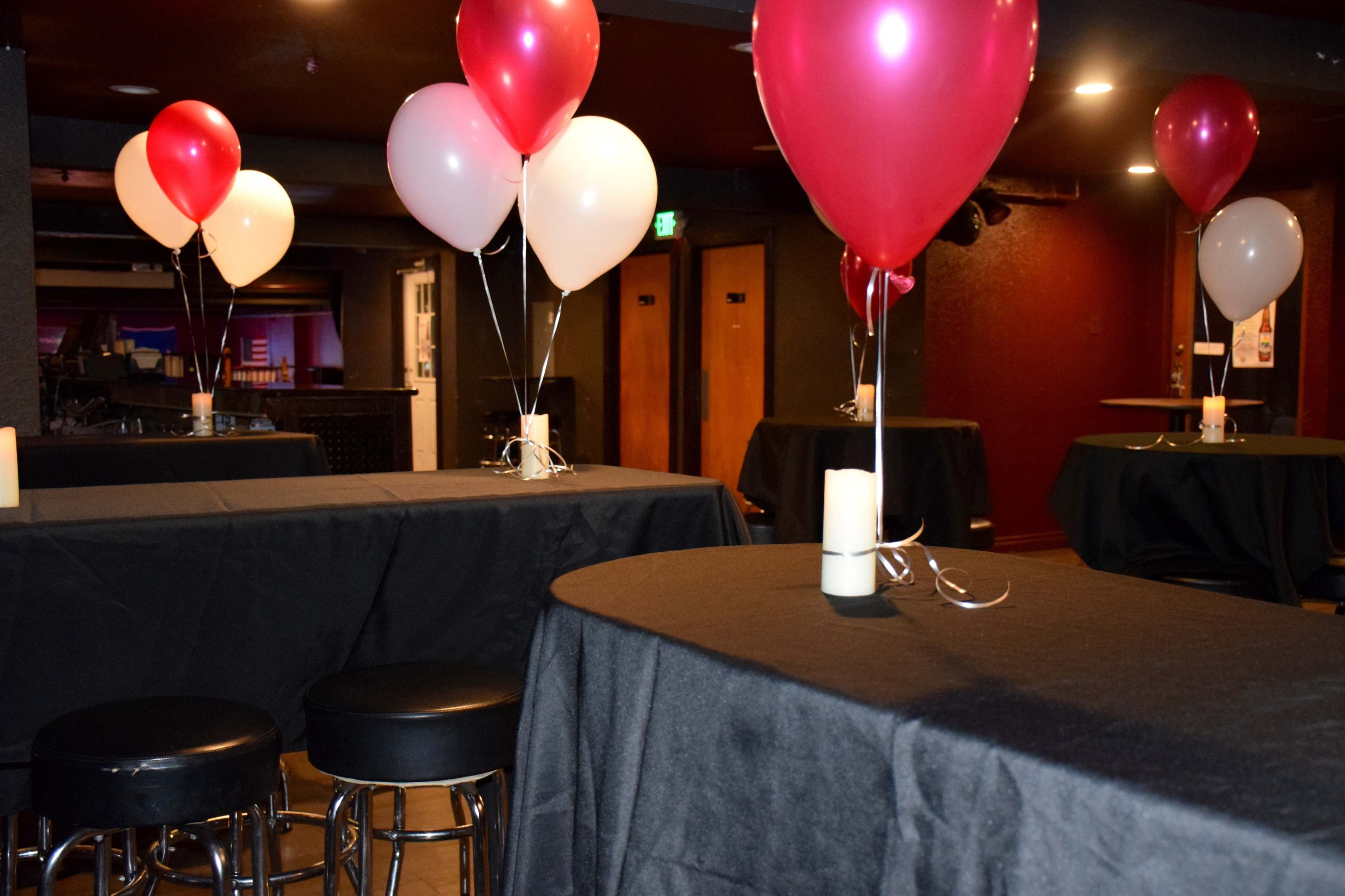 A dimly lit event space with black-covered tables, red and white balloons, and candles placed on the tables.