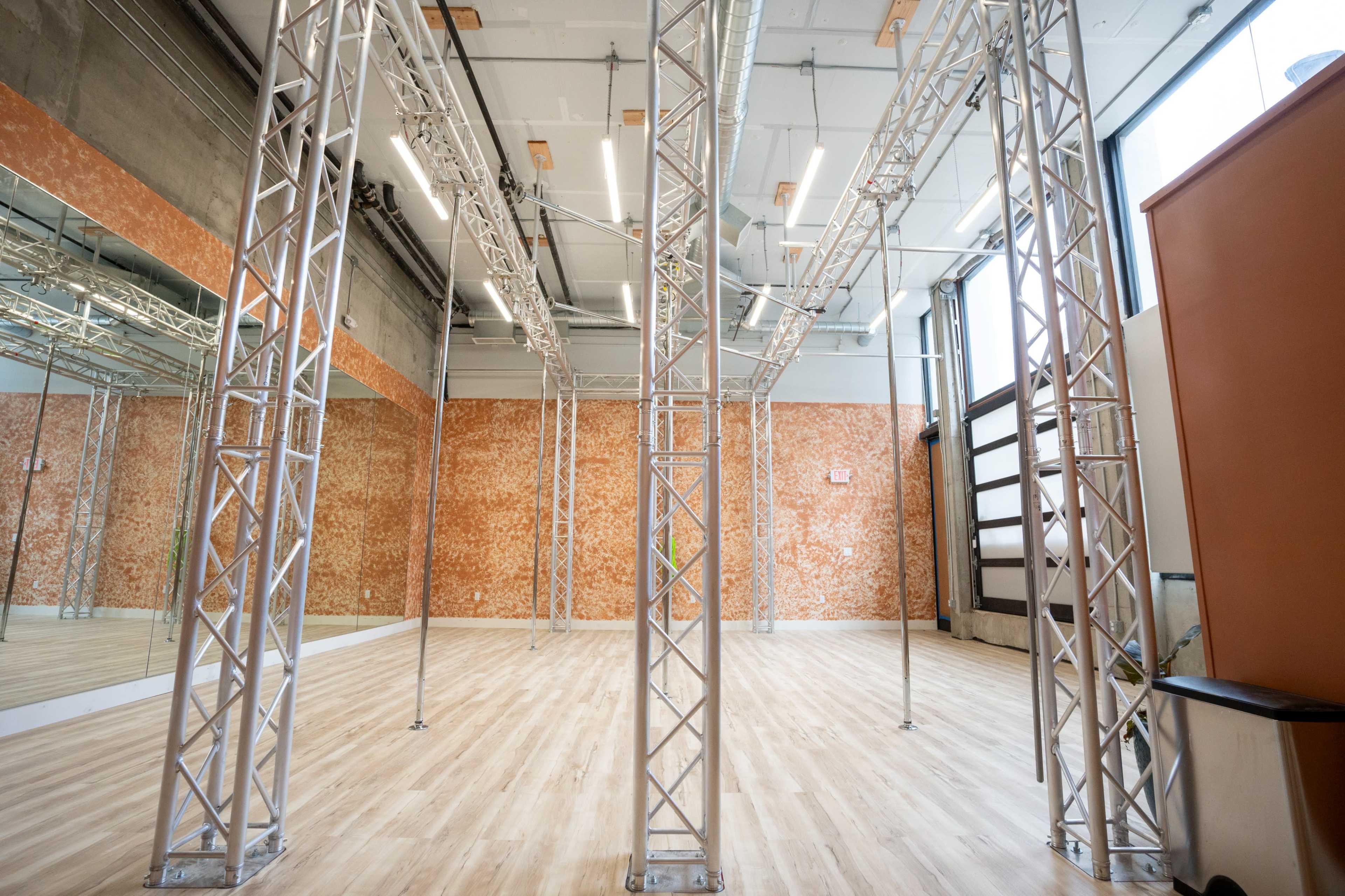 A spacious empty interior of a dance studio featuring metal trusses and wooden flooring with a mirrored wall.
