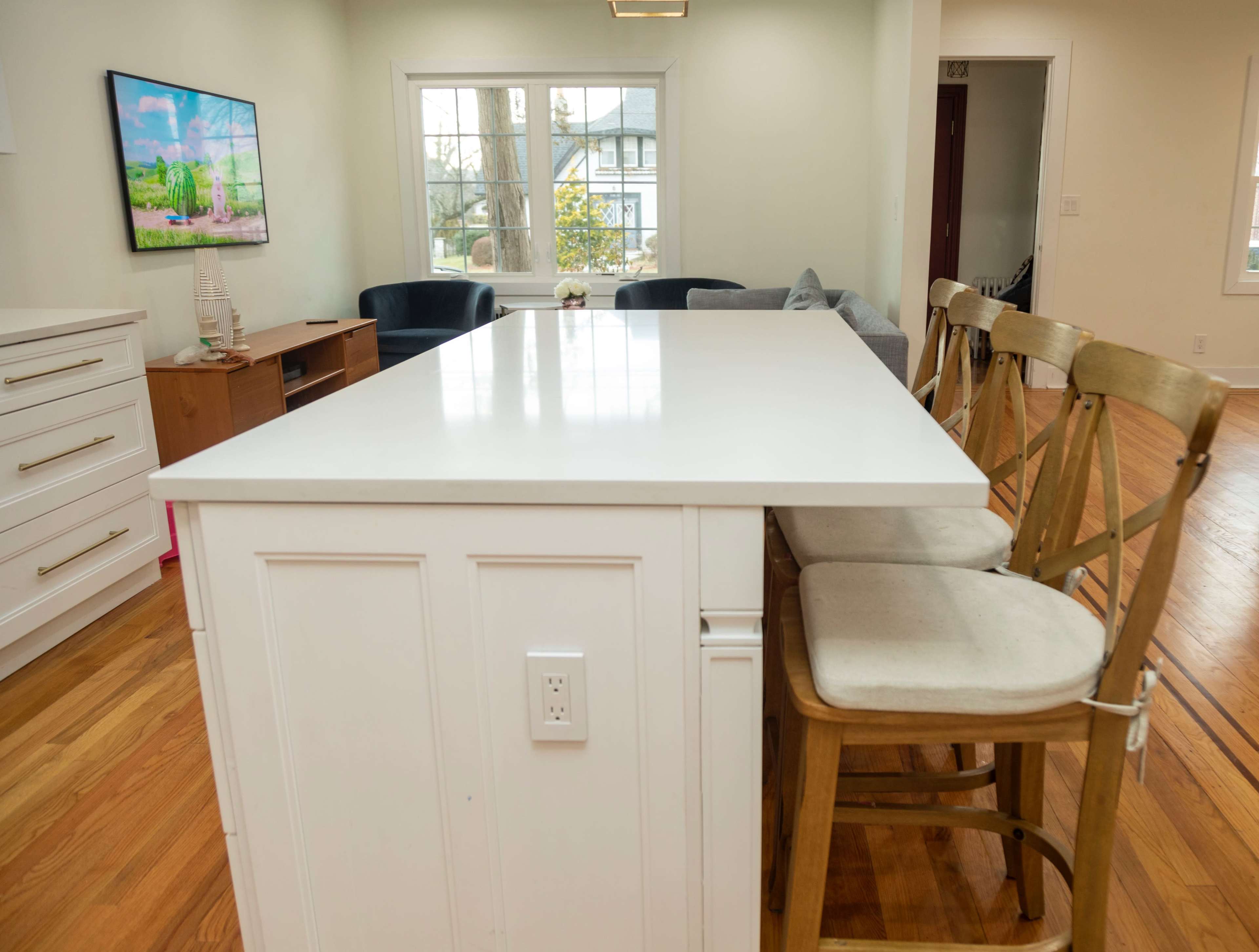 A kitchen island with four wooden stools is positioned in a bright, open-concept living area featuring a television and large windows.