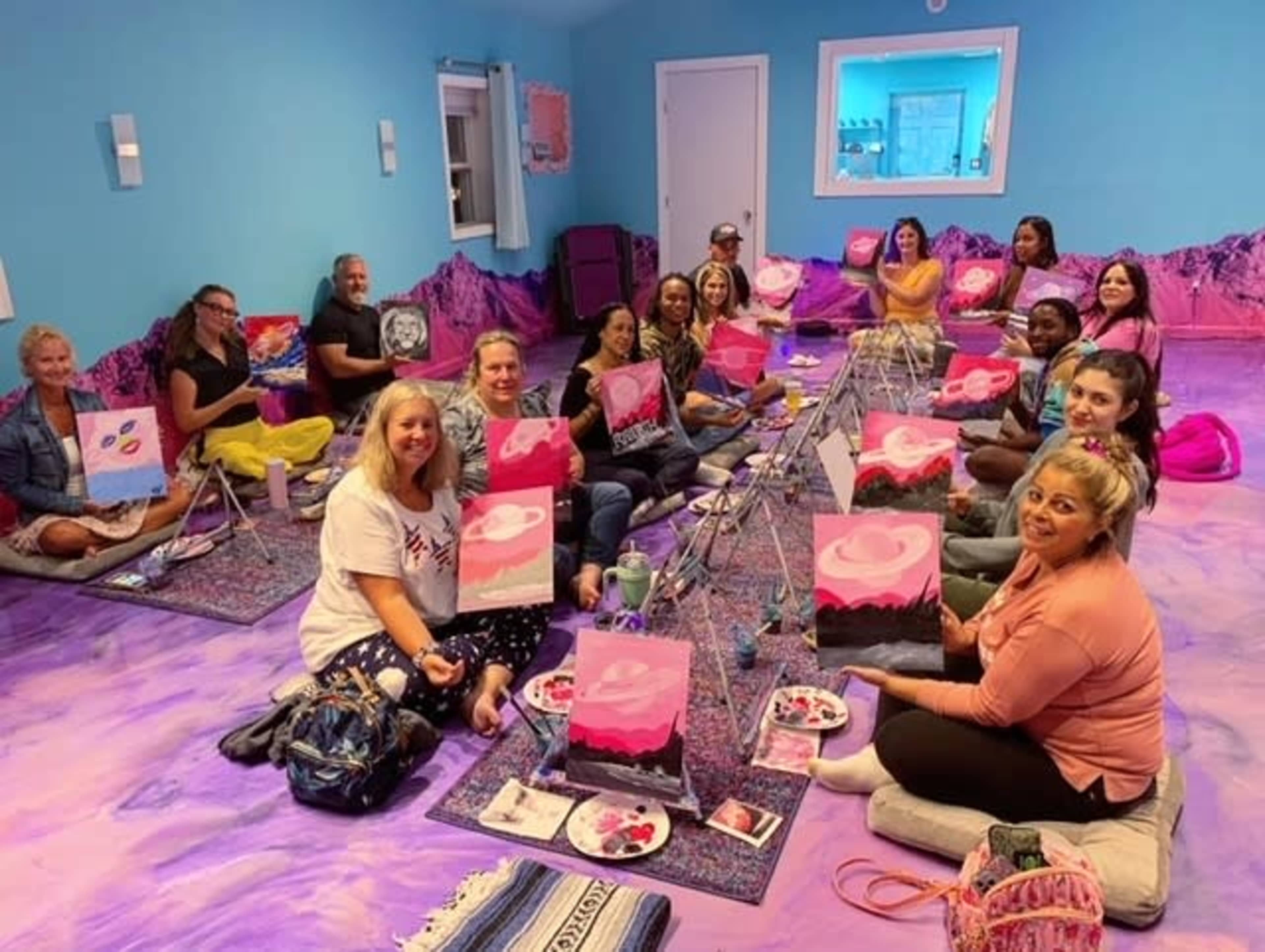 A group of people sit on the floor of a brightly colored room, each holding a painted canvas as they participate in a painting class.