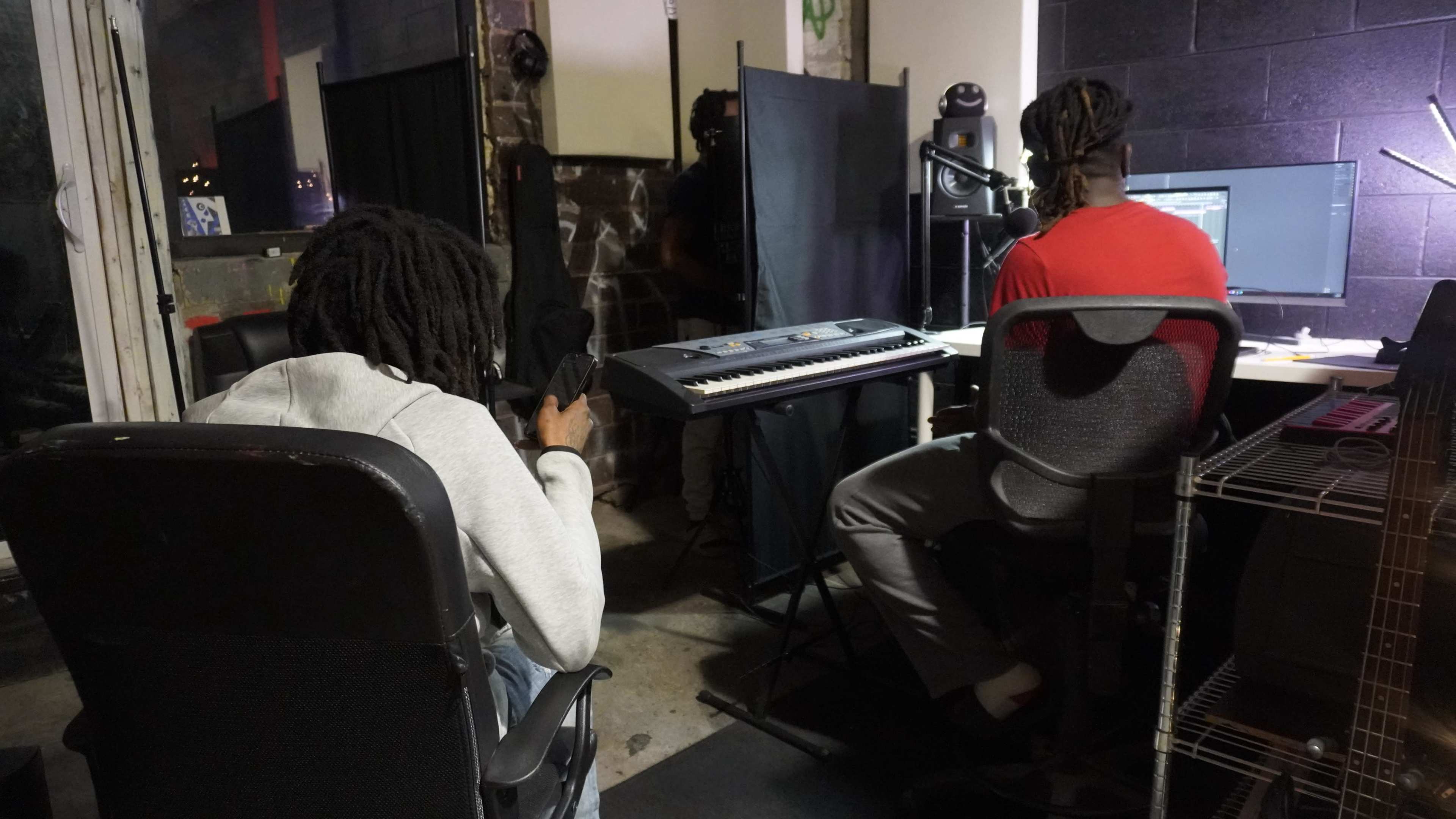 Two individuals are in a music studio, with one seated on a chair looking at their phone and the other at a computer with a keyboard in front of them.