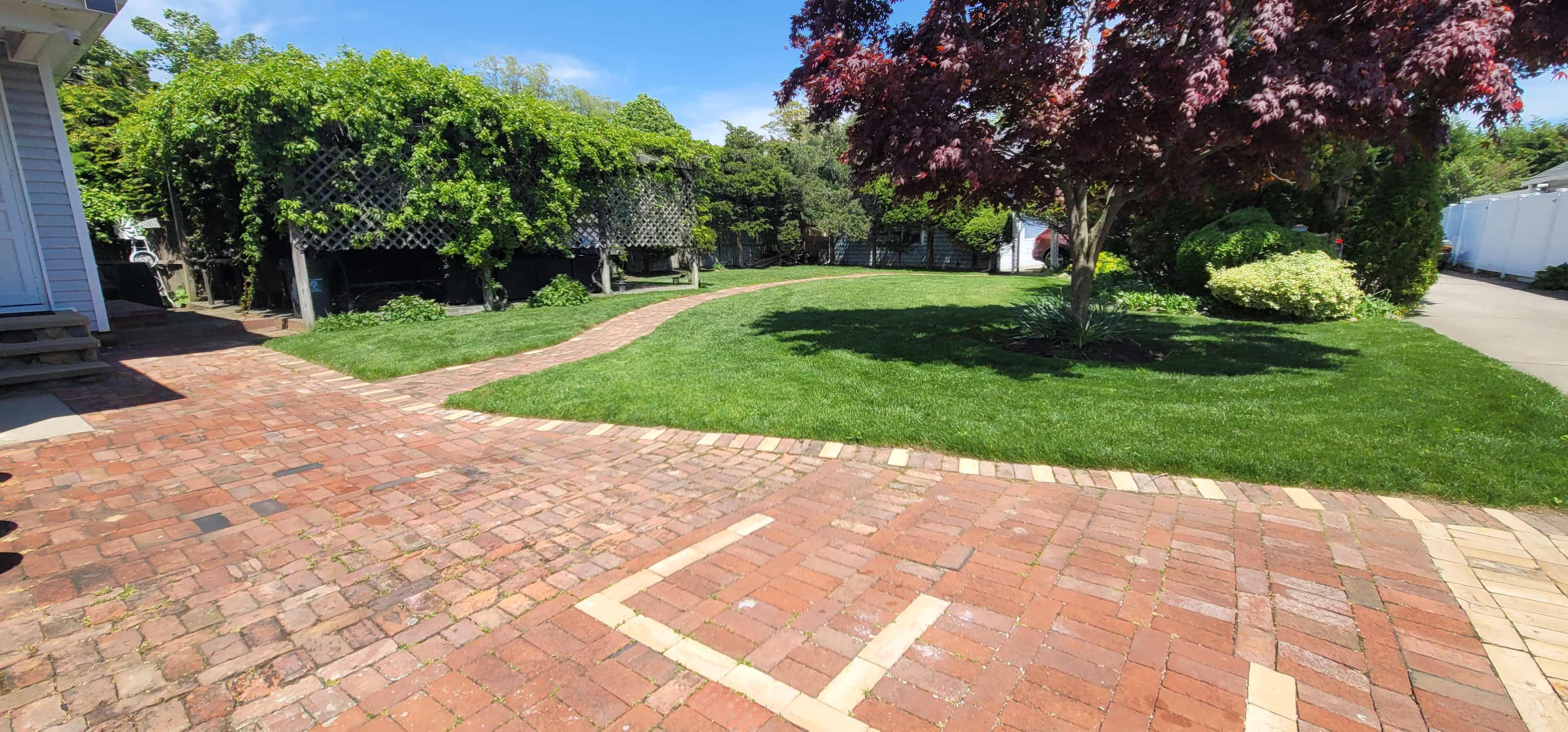 A brick pathway winds through a manicured green lawn surrounded by trees and shrubs in a residential backyard.