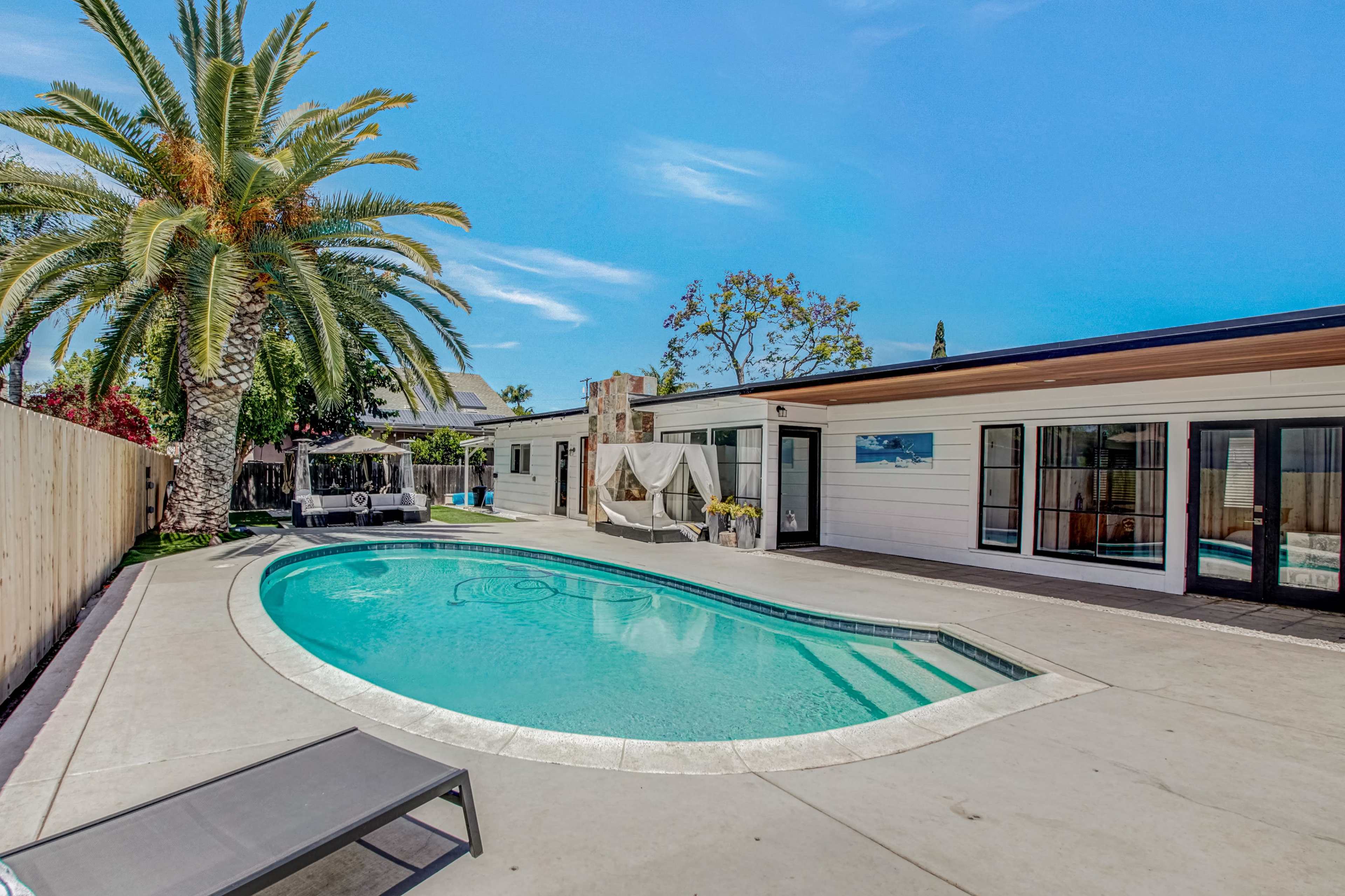 A swimming pool surrounded by a concrete patio, with palm trees and a house featuring large glass doors in the background.