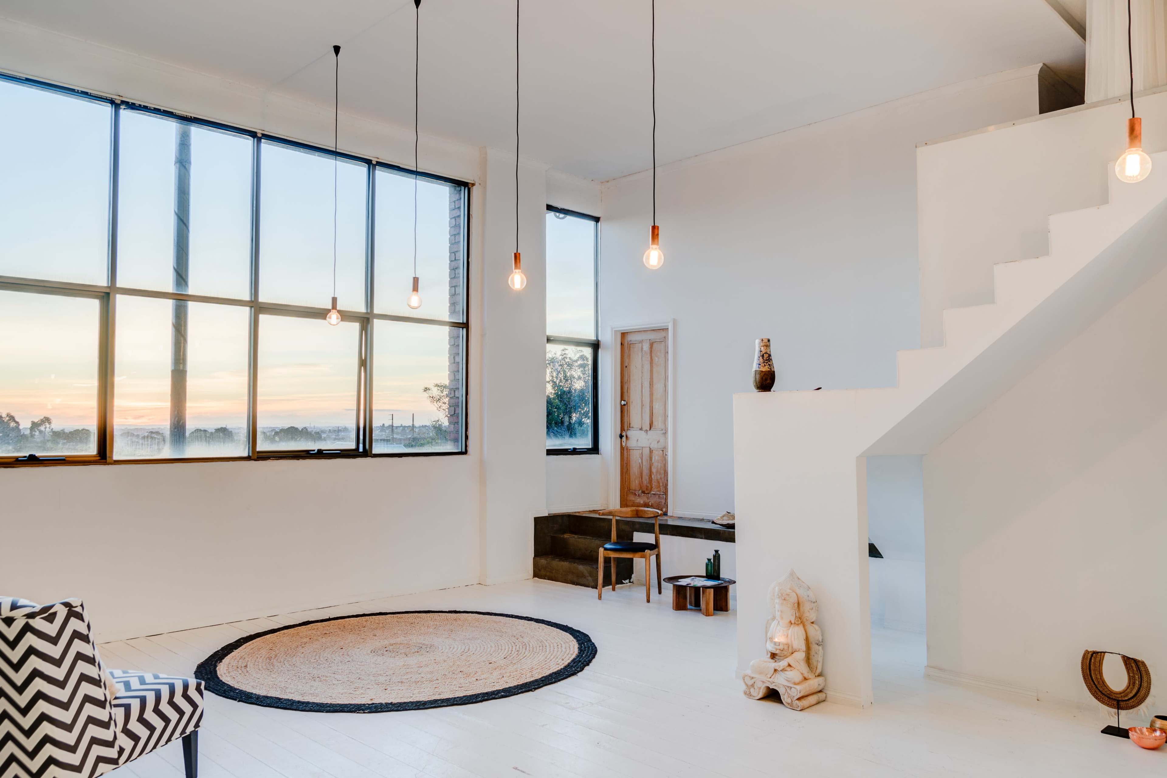 The room features large windows that provide a view of the sunset, a staircase leading to an upper level, and minimalistic furnishings including a round rug and a decorative statue.