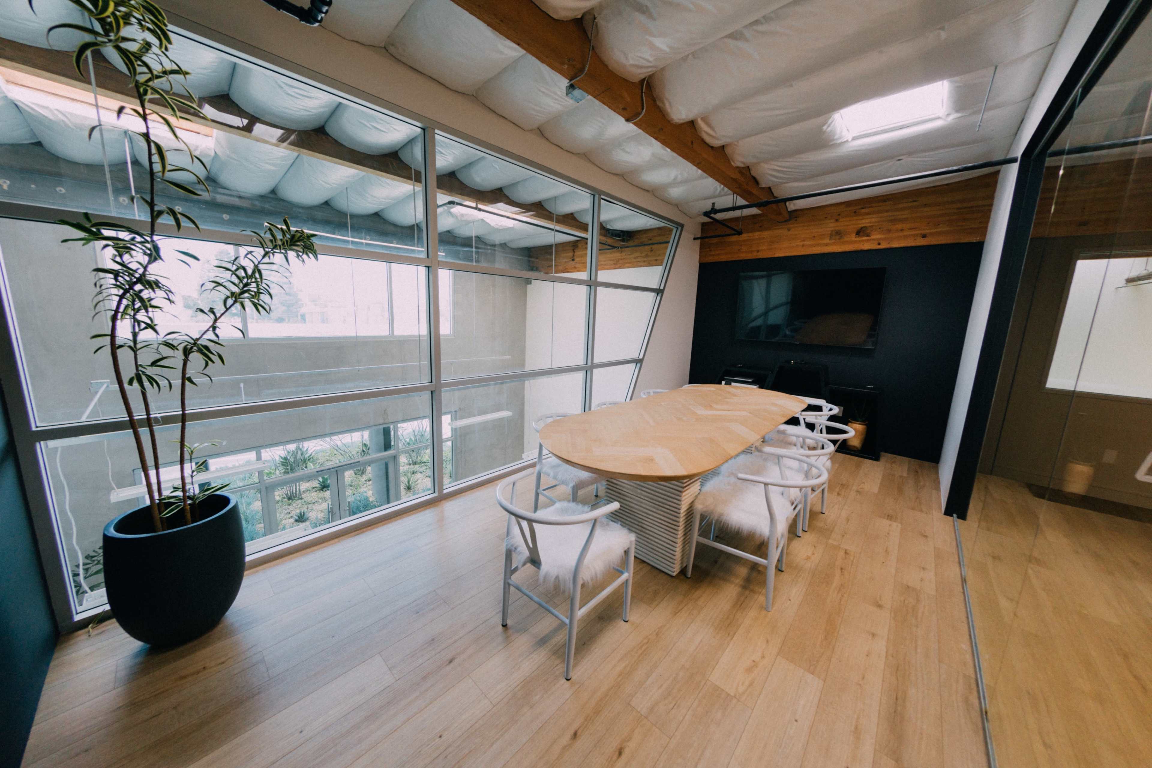 A modern conference room features a large, wooden table surrounded by white chairs, with large windows letting in natural light.