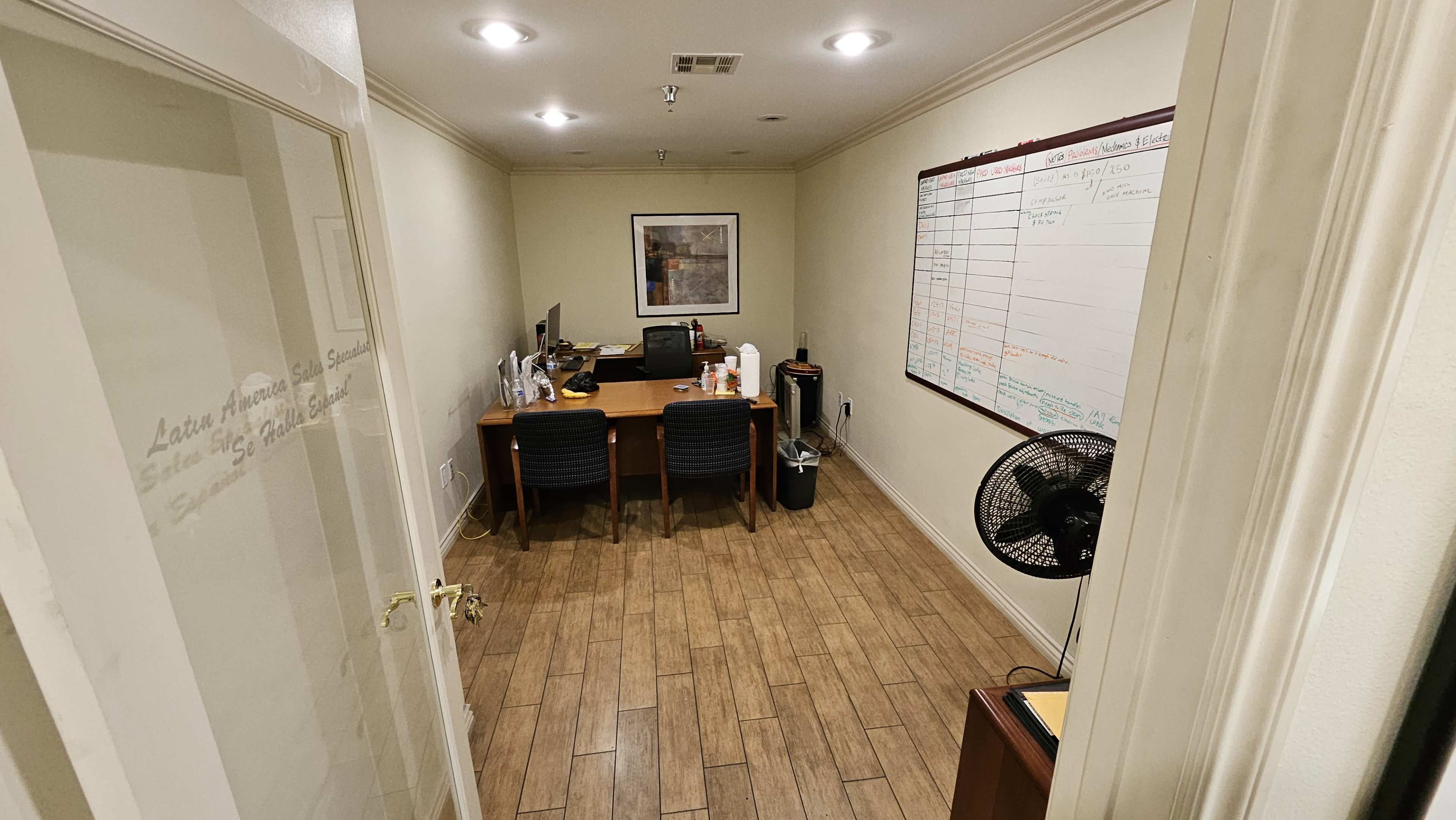 A small office space features a desk with a computer, chairs, a fan, and a whiteboard on the wall.