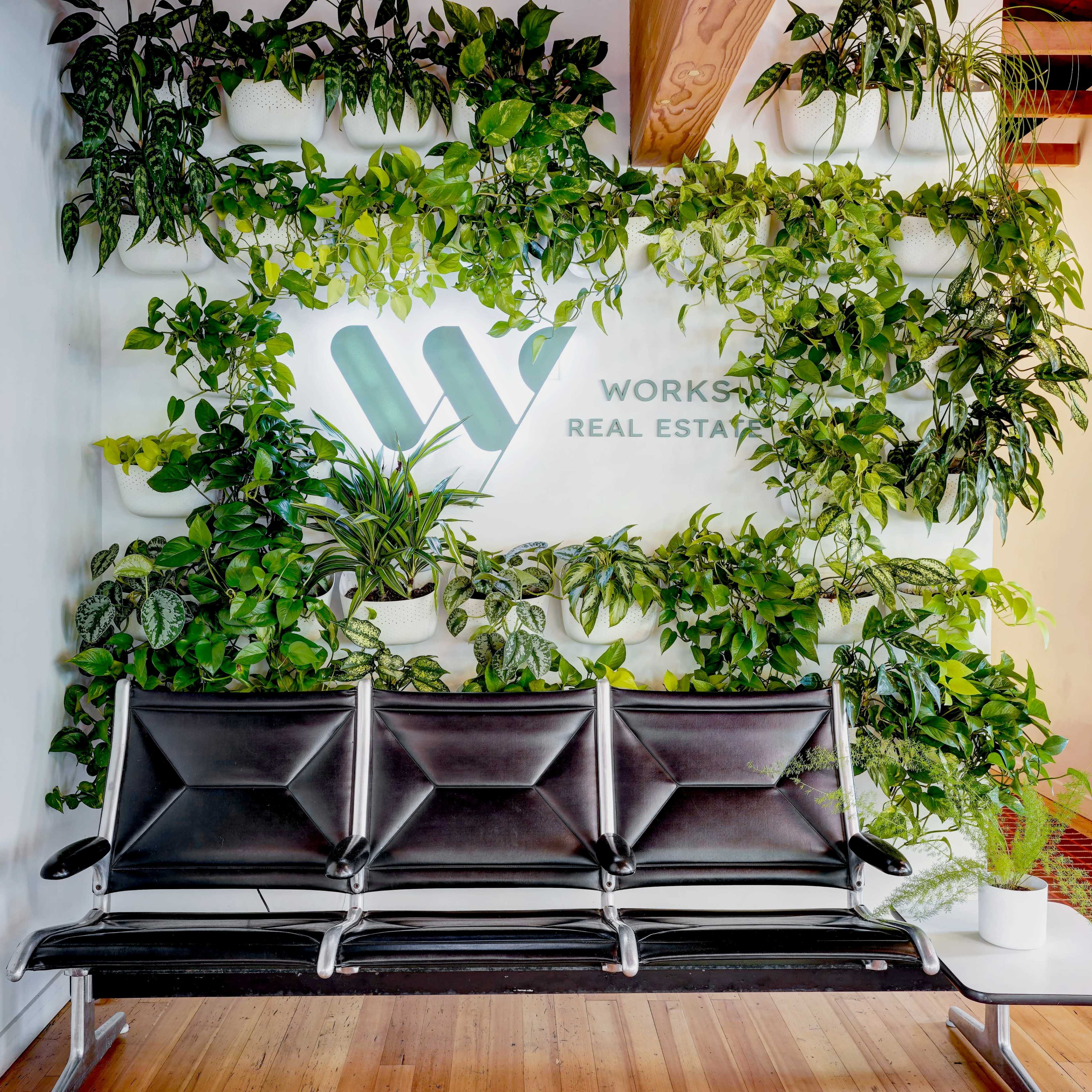 A black three-seat bench is positioned in front of a wall adorned with various green plants and a logo that reads "WORKSPACE REAL ESTATE."