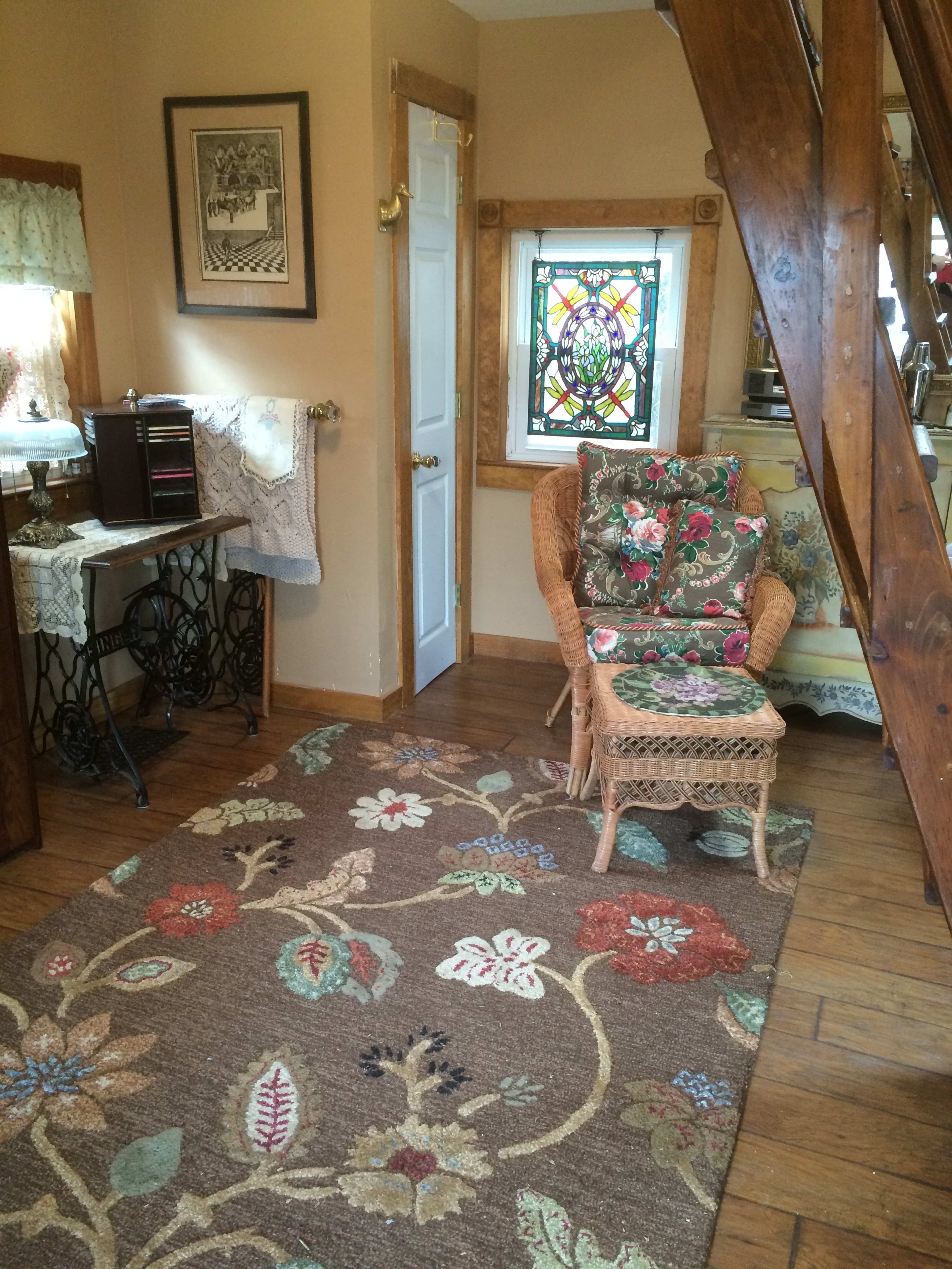 The image shows a cozy corner of a room featuring a wicker chair with floral cushions, a patterned rug, and a decorative stained glass window.