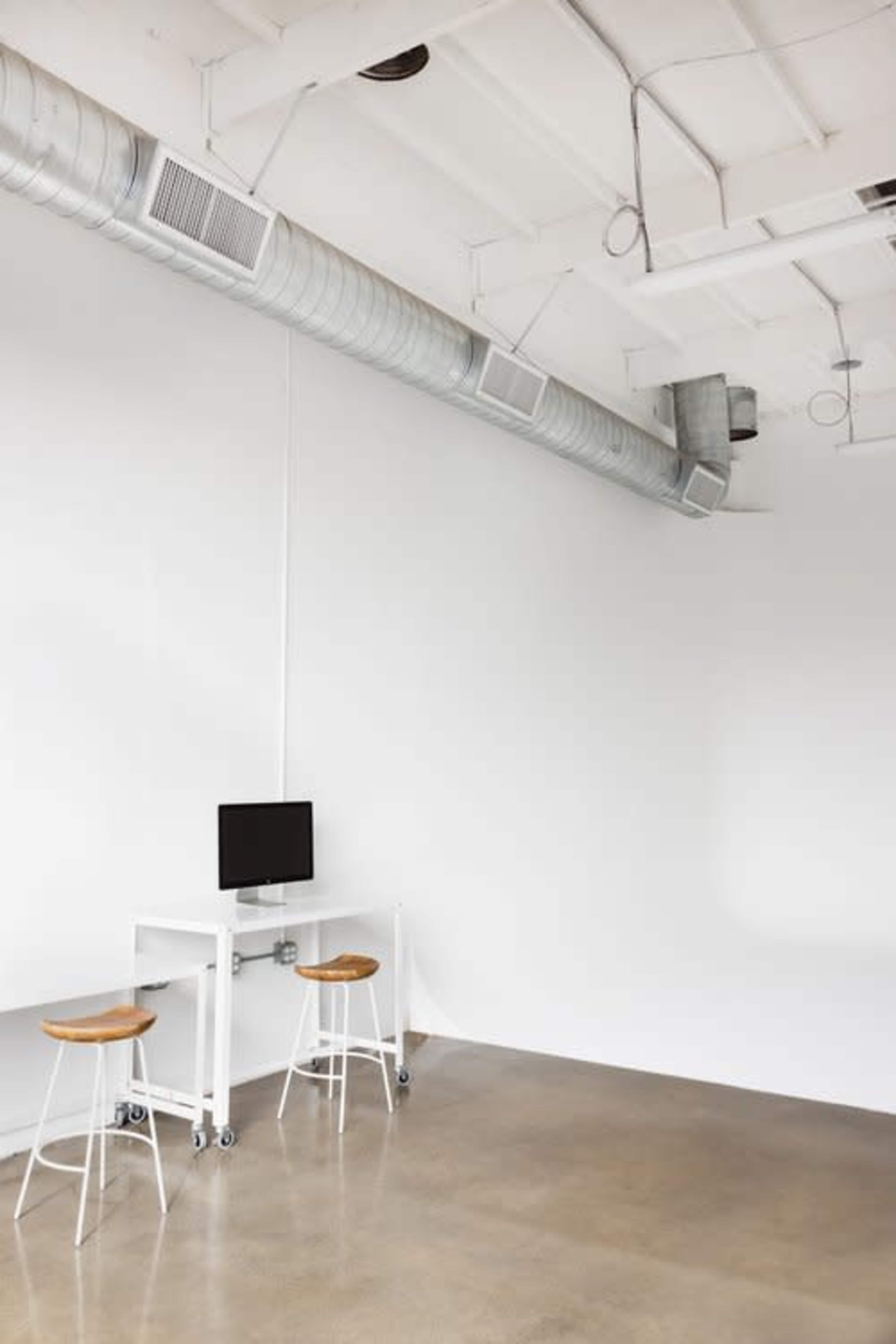 A minimalistic room with a metal desk on wheels, a computer monitor, and two wooden stools against a plain white wall.