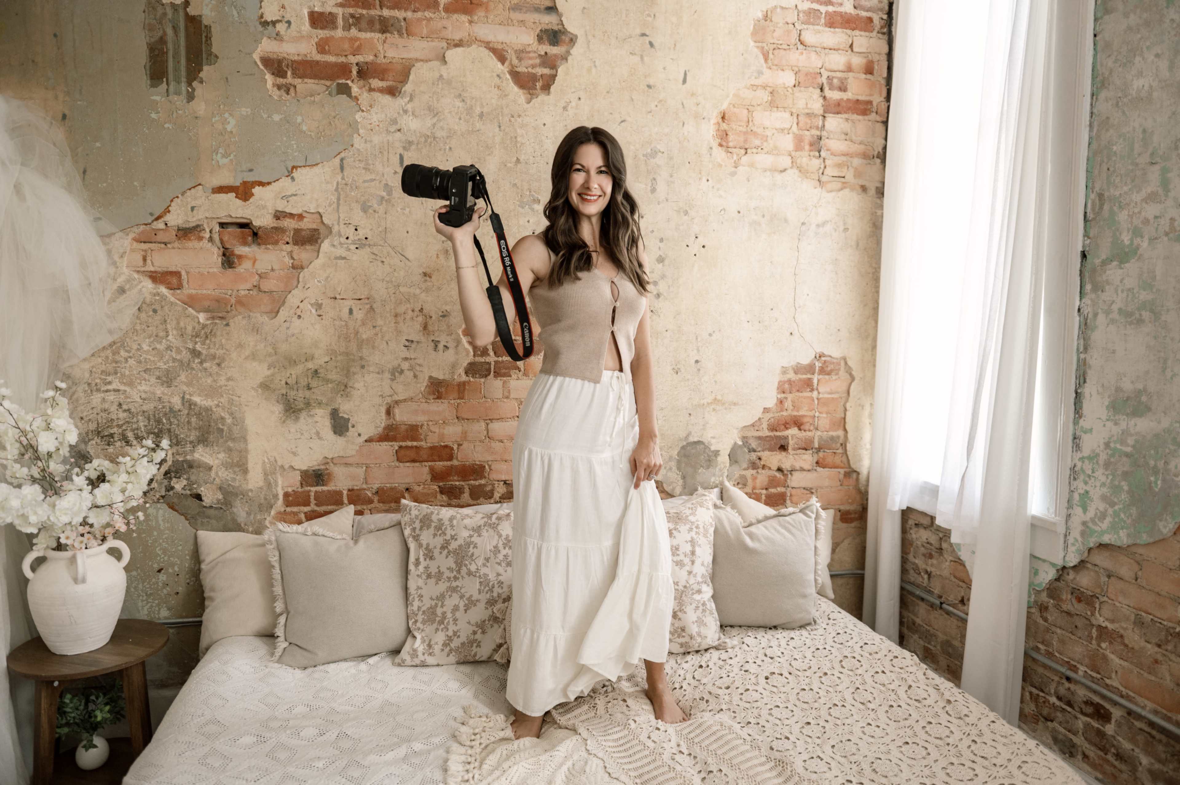 A woman stands on a bed in a rustic room with exposed brick walls, holding a camera and wearing a light-colored outfit.