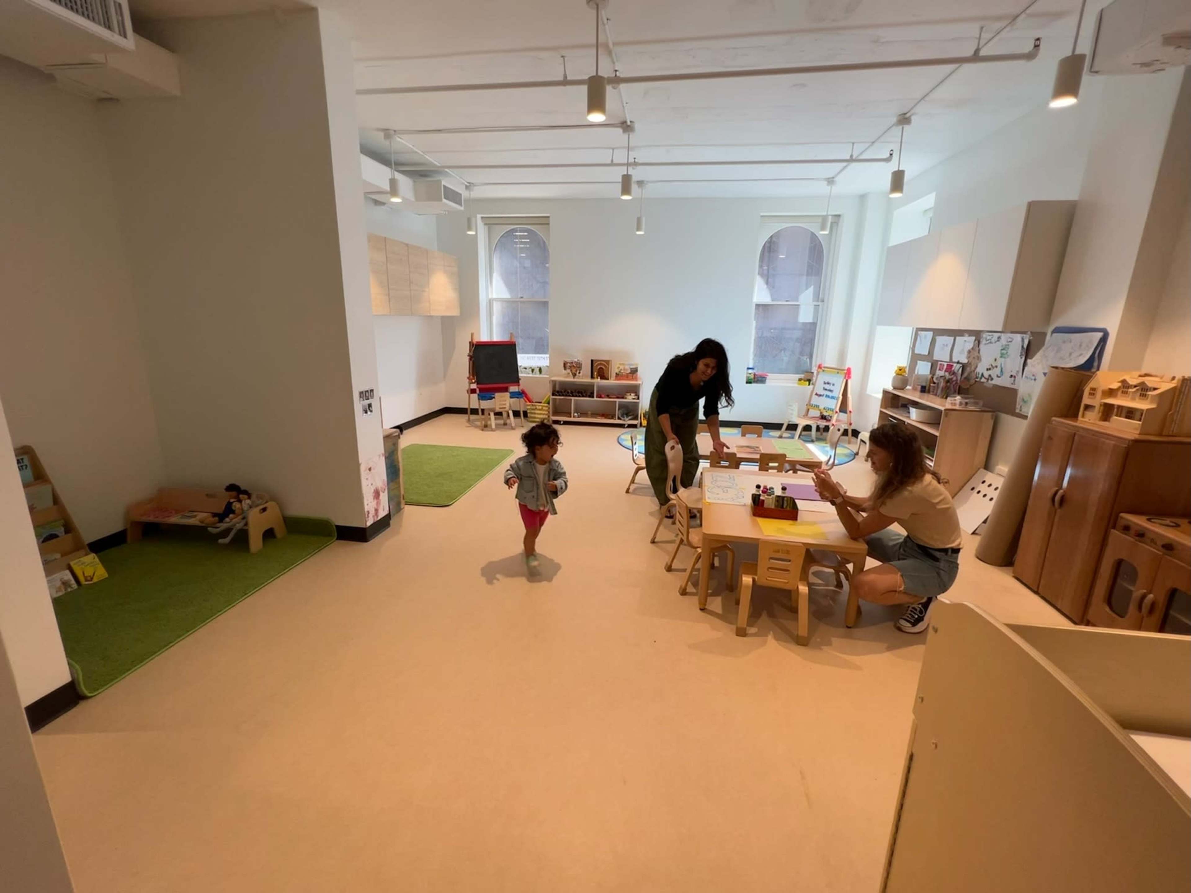 A child runs toward a table where two adults are engaged in an activity in a bright, spacious room designed for play and learning.
