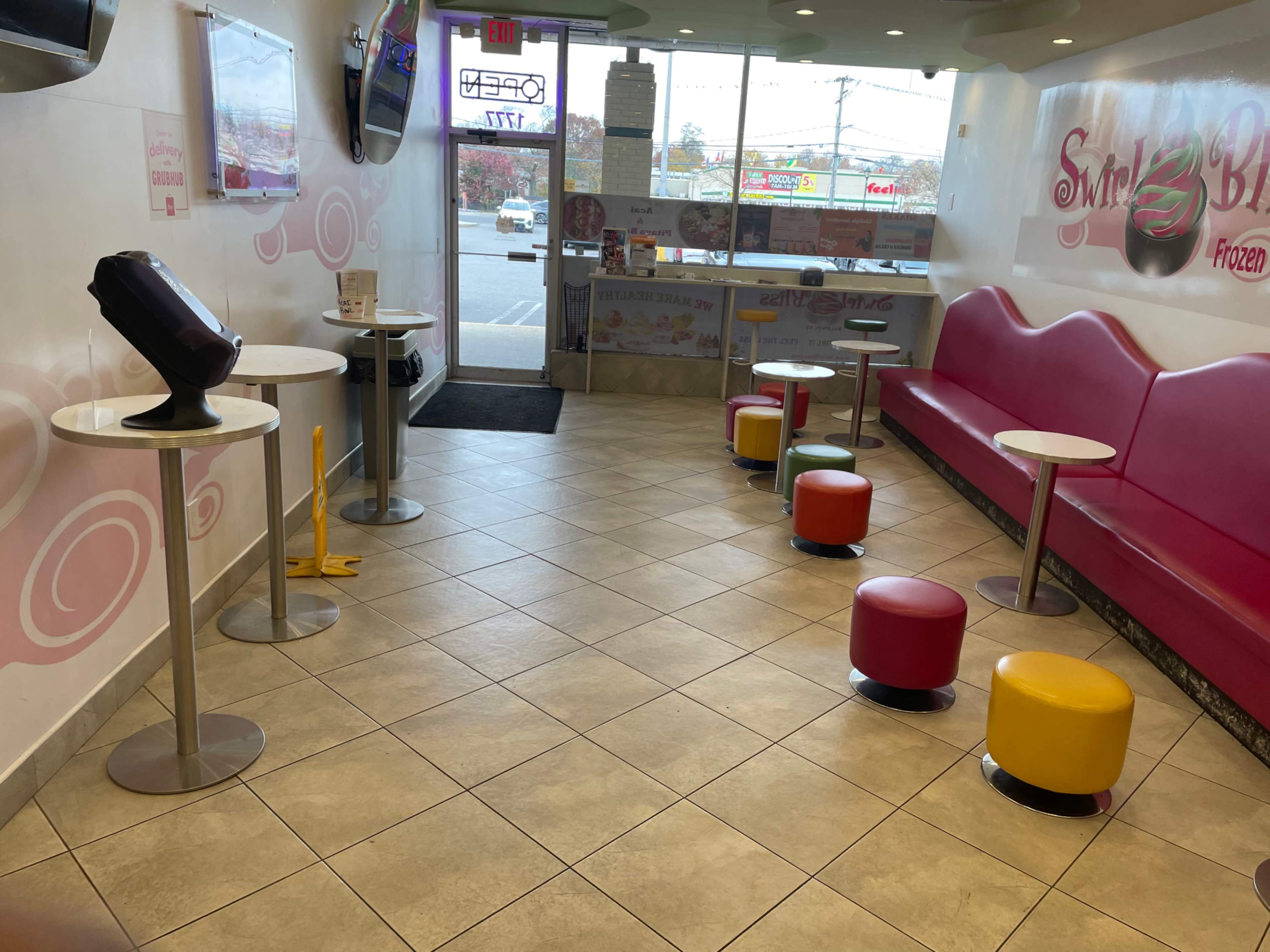 The interior of a frozen yogurt shop features a pink seating area, circular tables, and colorful stools, with a view of the entrance and outdoor area.