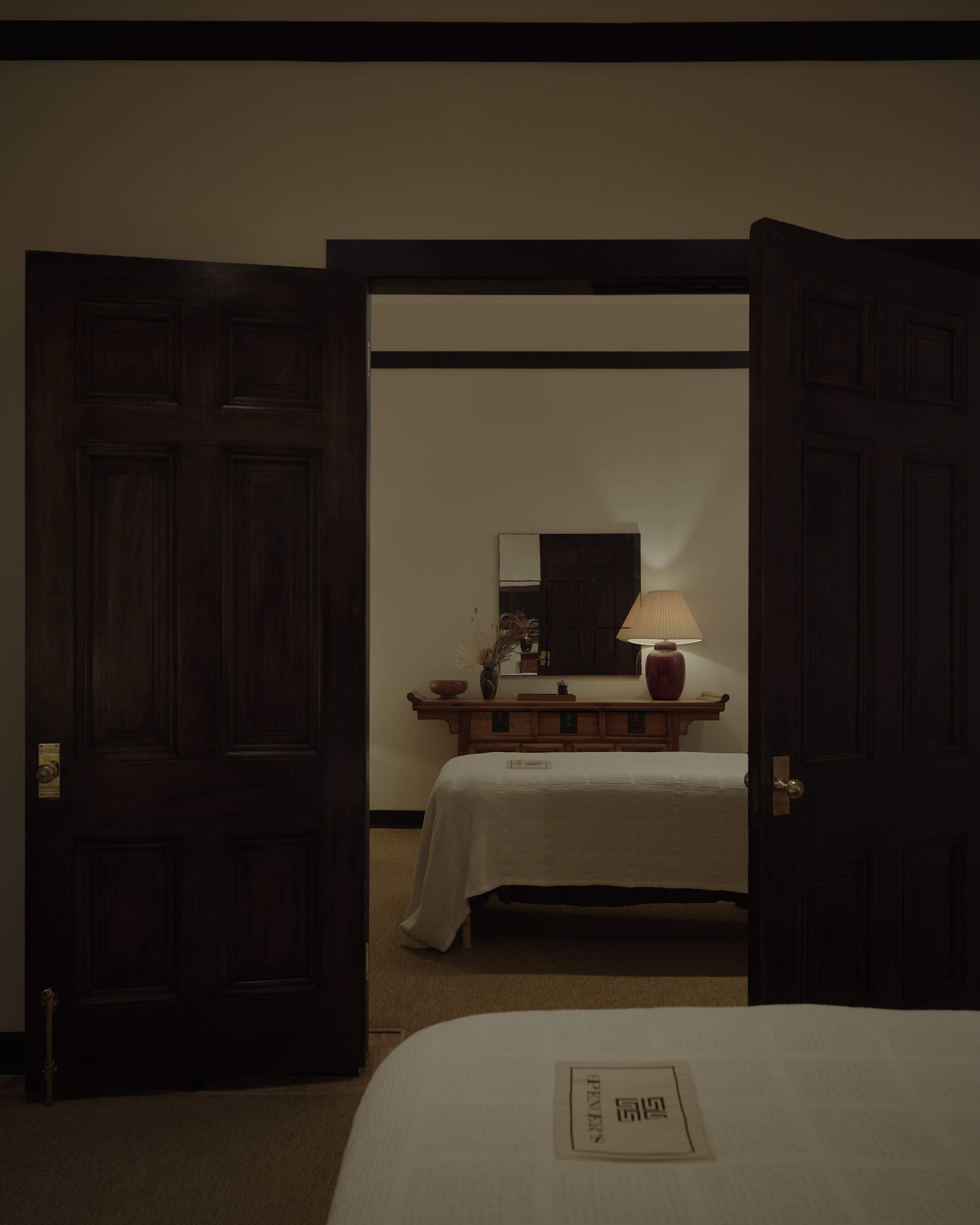 A lighted bedroom is visible through an open doorway, featuring a dresser with a mirror, a lamp, and a bed with a white coverlet.