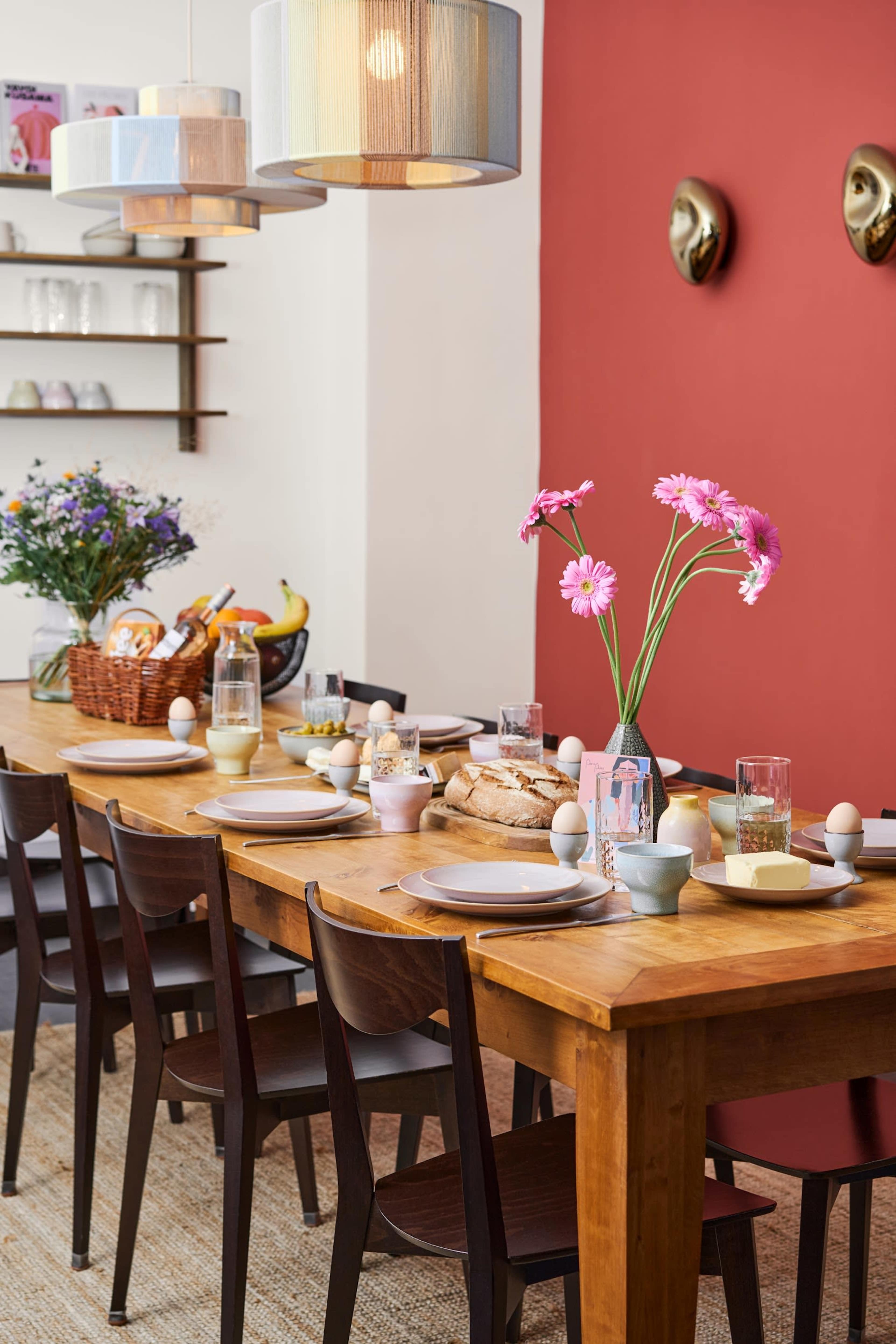 A dining table is set with plates, glasses, and vases of flowers, surrounded by chairs in a brightly colored room.