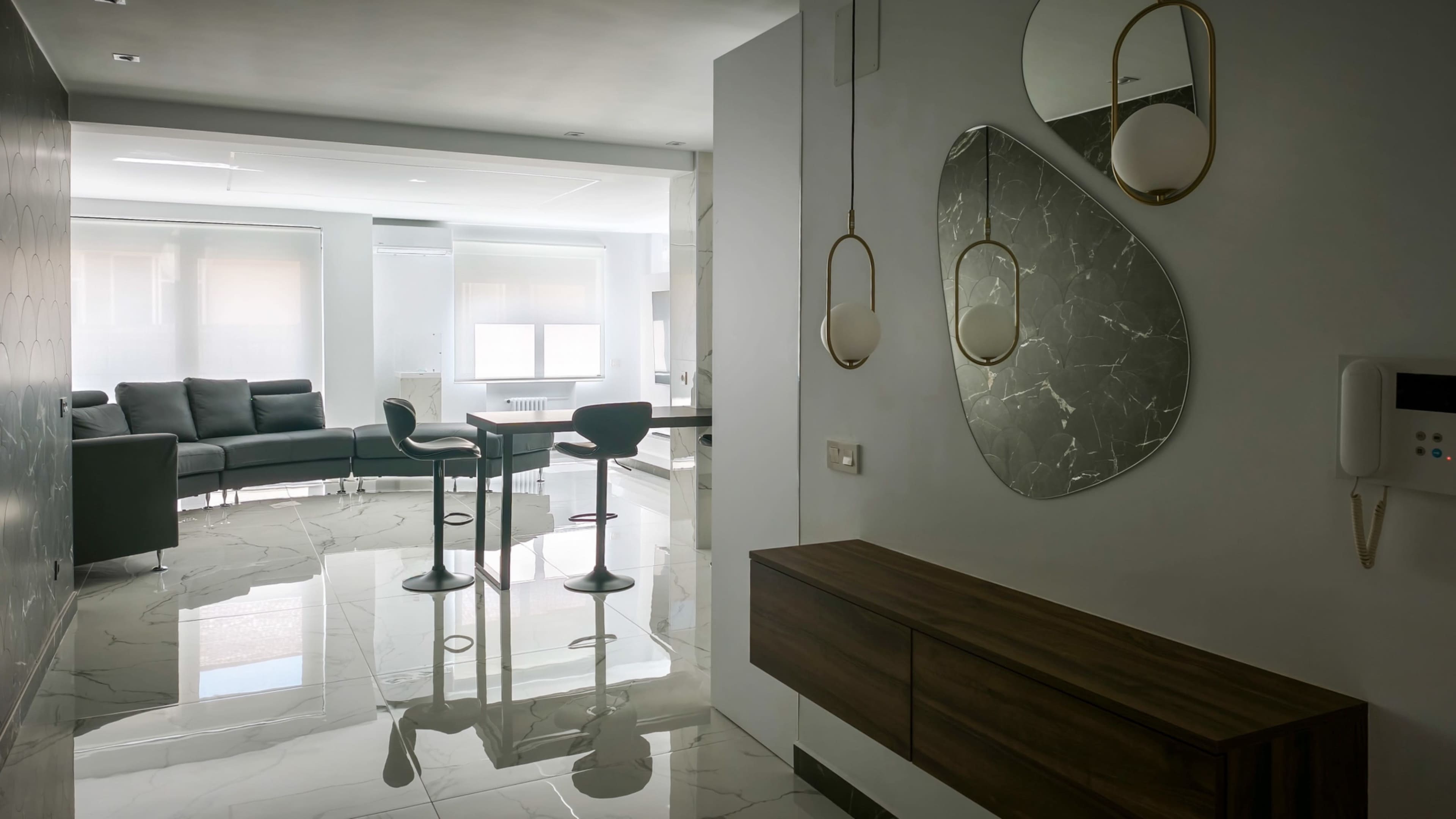 The image shows a modern interior space featuring a hallway with a console table and mirrors, leading to a living area with a sofa and bar stools.