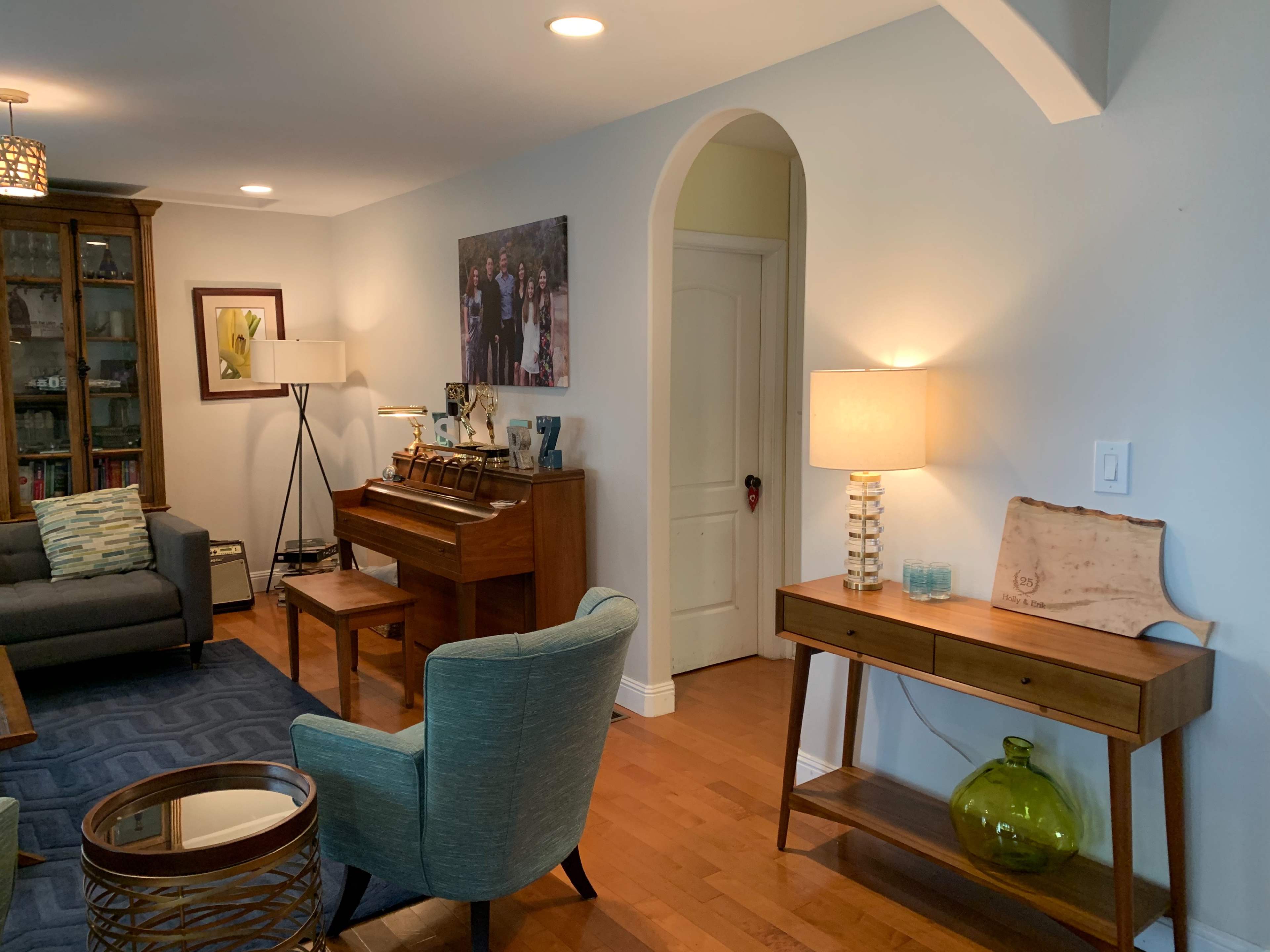 A living room features a wooden piano, a table lamp, blue accent chairs, and a decorative console table against light-colored walls.