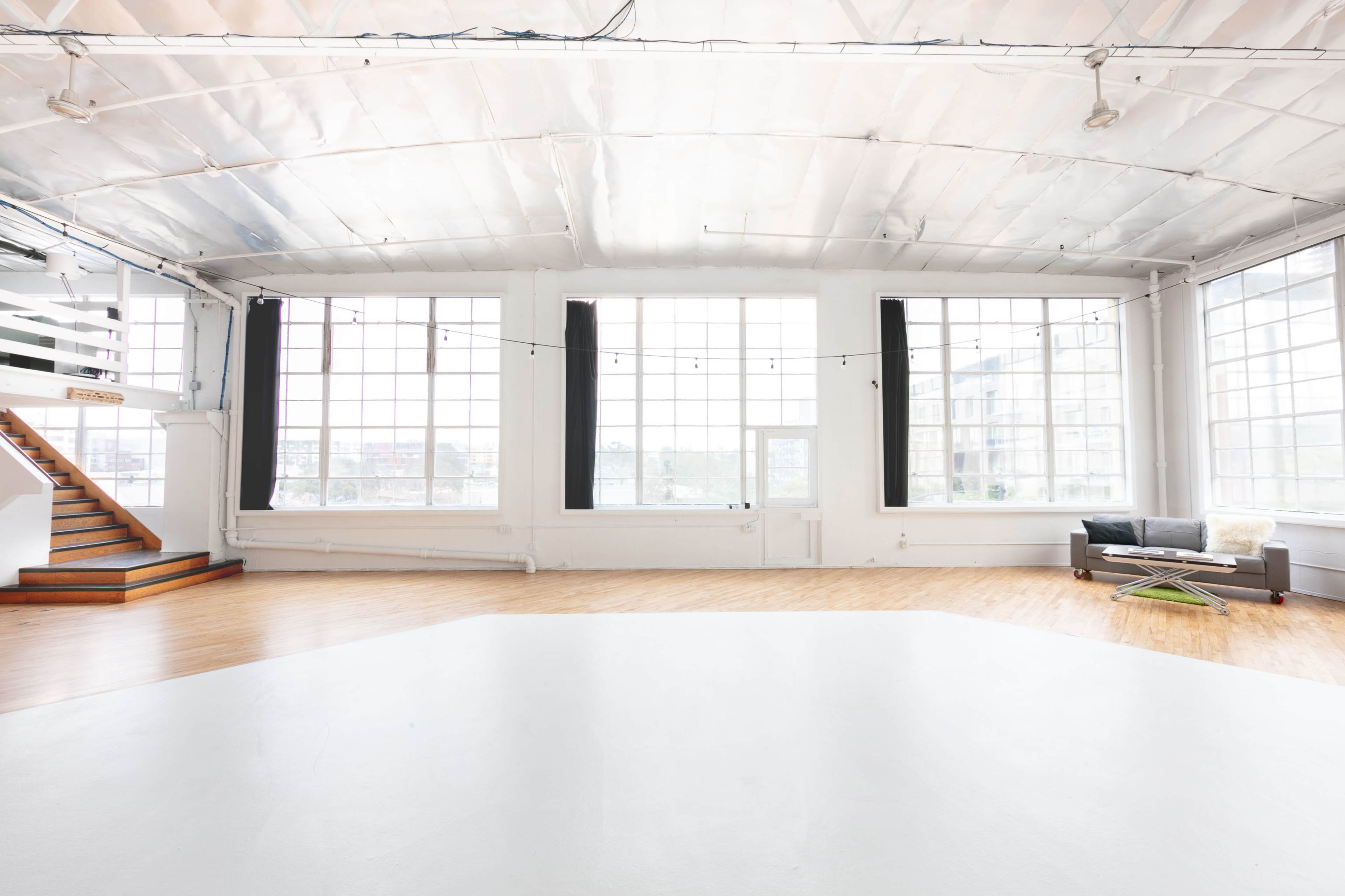 The image shows a spacious, bright interior with large windows, wooden flooring, and a staircase leading to an upper level.