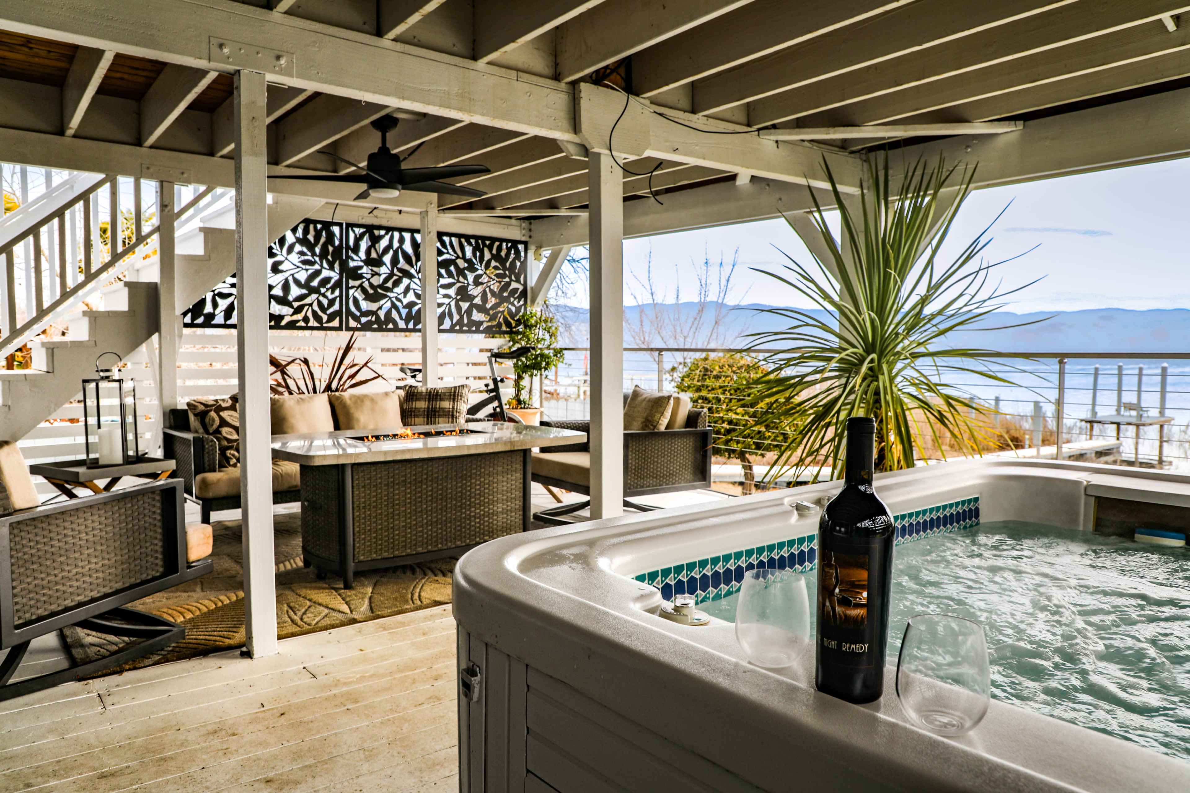 The scene features a covered outdoor seating area with a hot tub, a dining table, and views of a lake and mountains in the background.