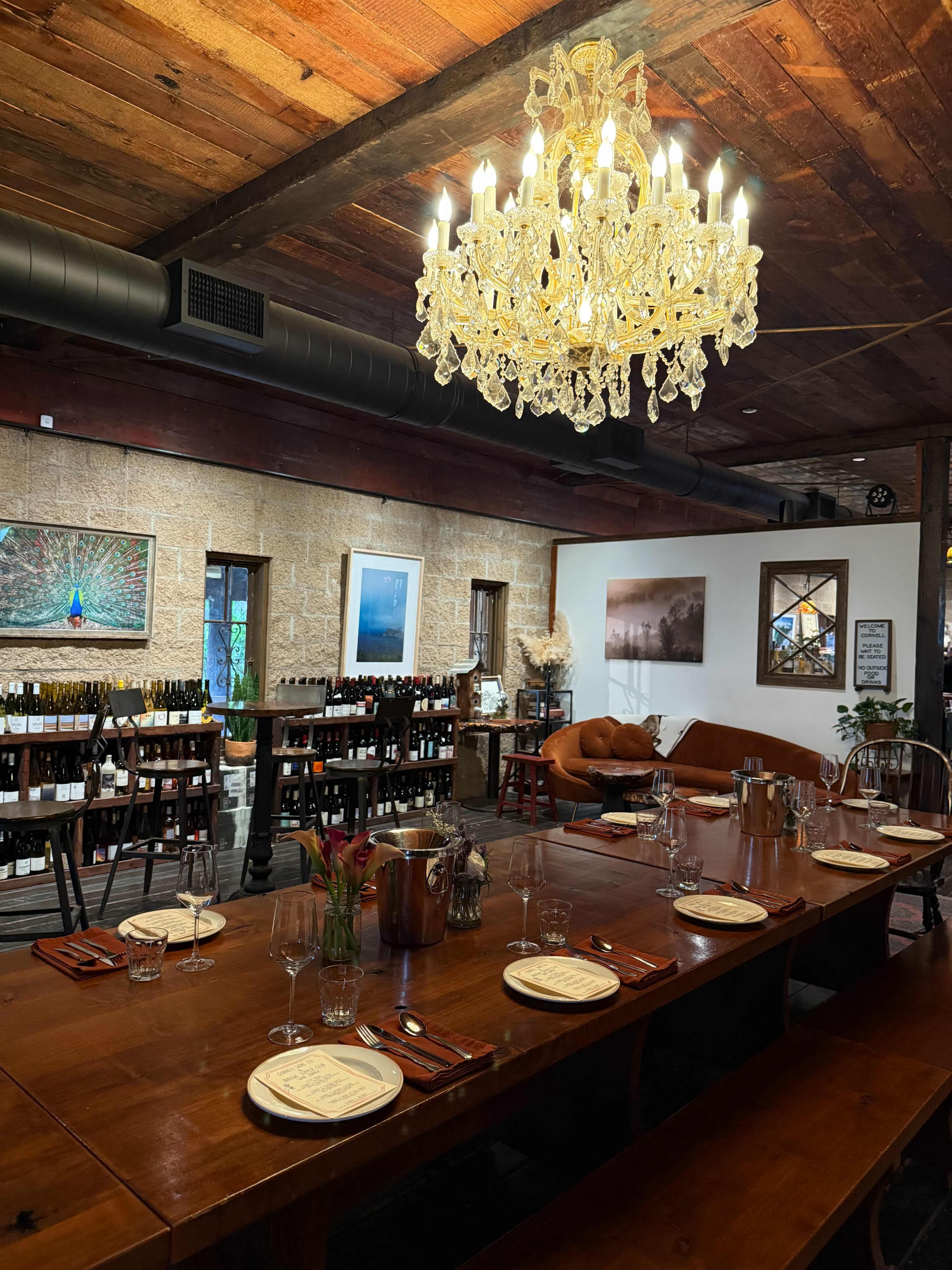 The image shows a rustic dining area with a long wooden table set for a meal, surrounded by wine bottles and adorned with a large chandelier.