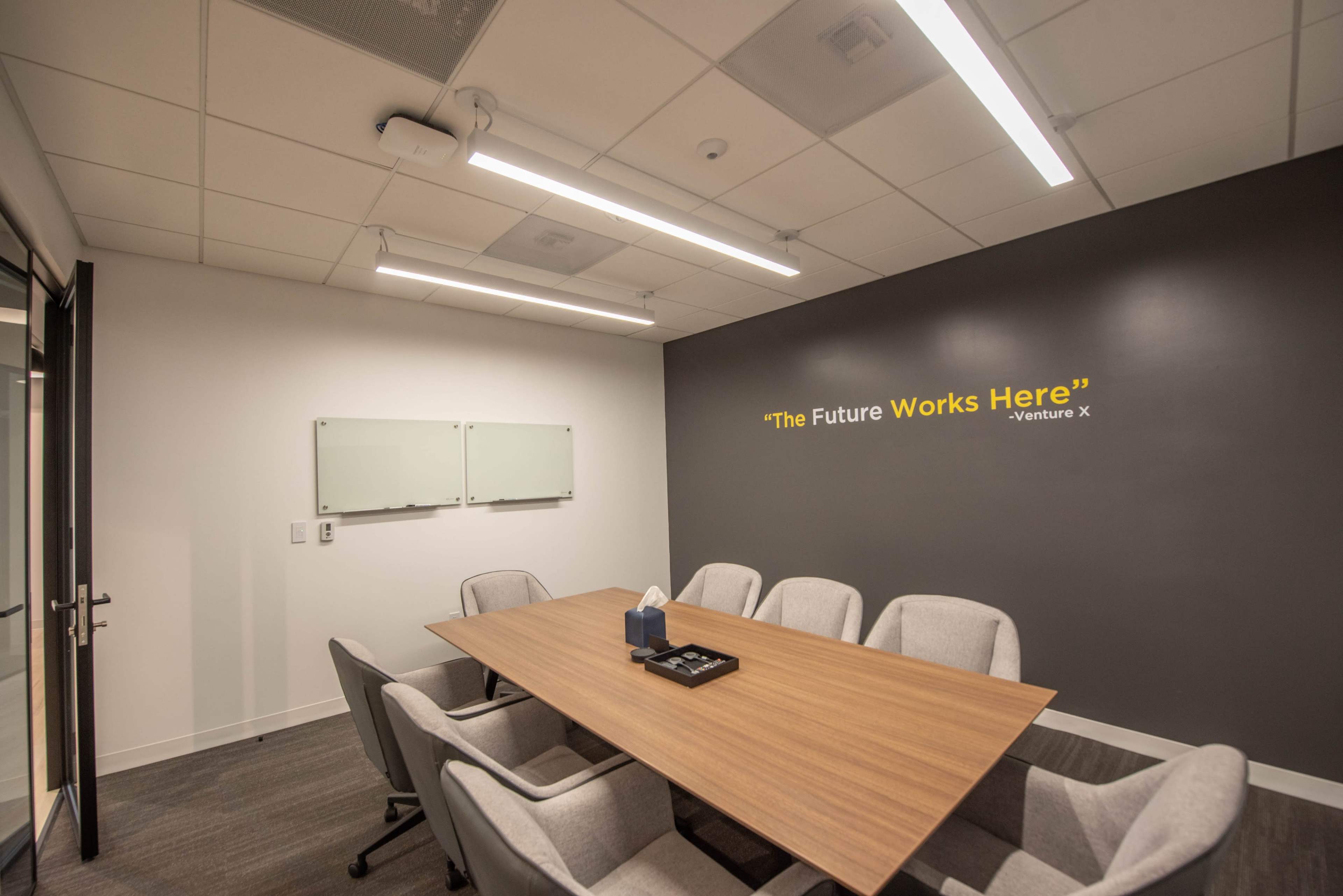 A modern conference room features a wooden table surrounded by six gray chairs and a wall with a motivational quote.