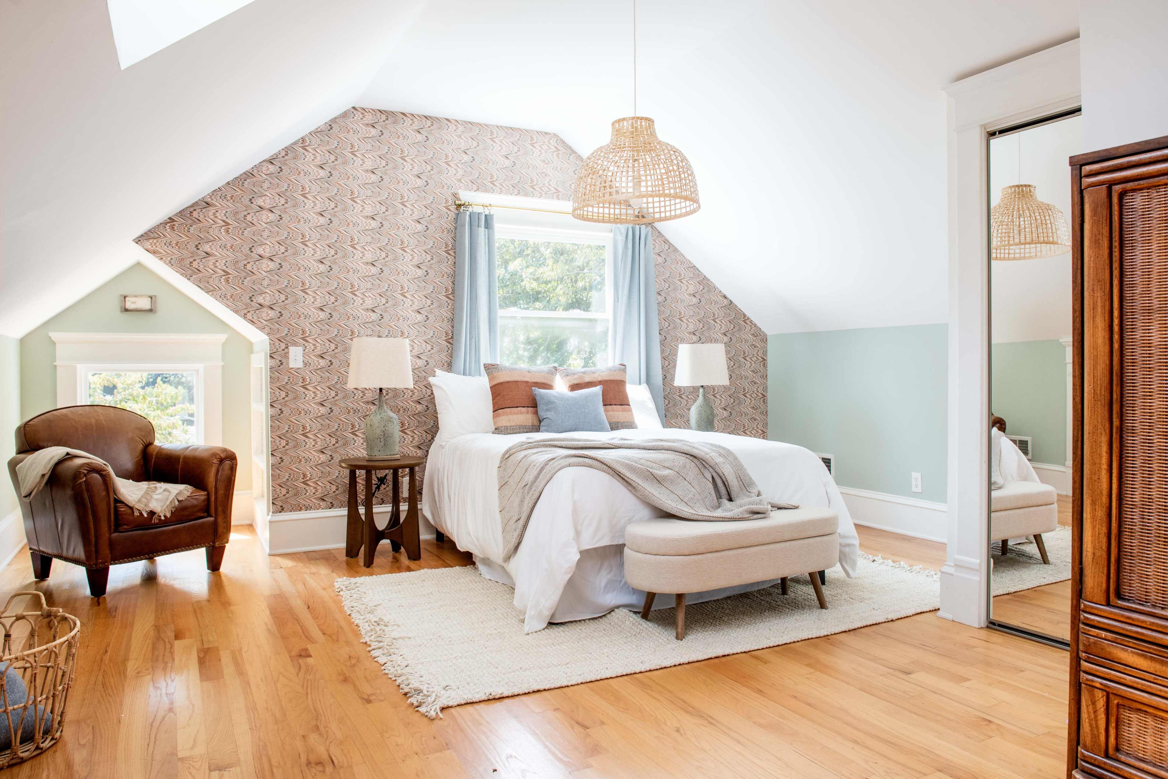 A spacious, well-lit bedroom features a bed with light bedding, a cozy armchair, and wooden flooring, complemented by patterned wallpaper and a mirror.