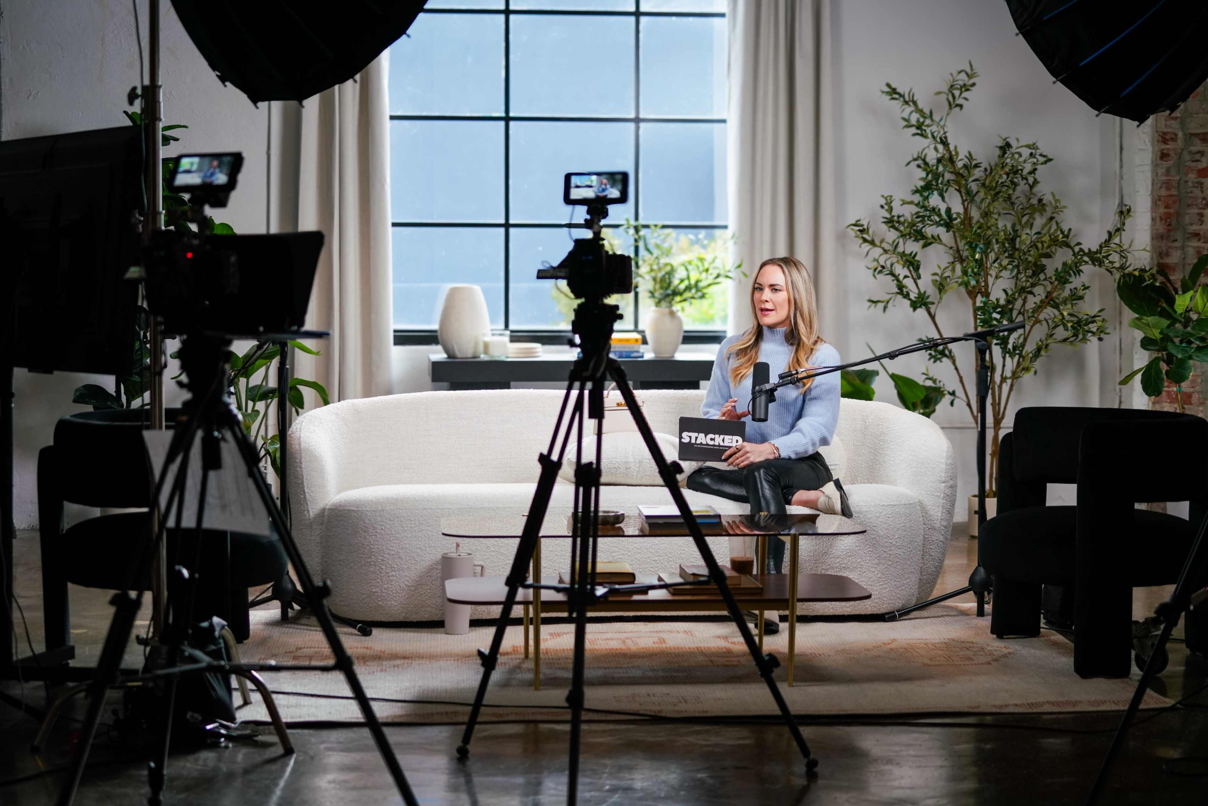 A woman sits on a white couch in a well-lit studio, flanked by cameras and plants.