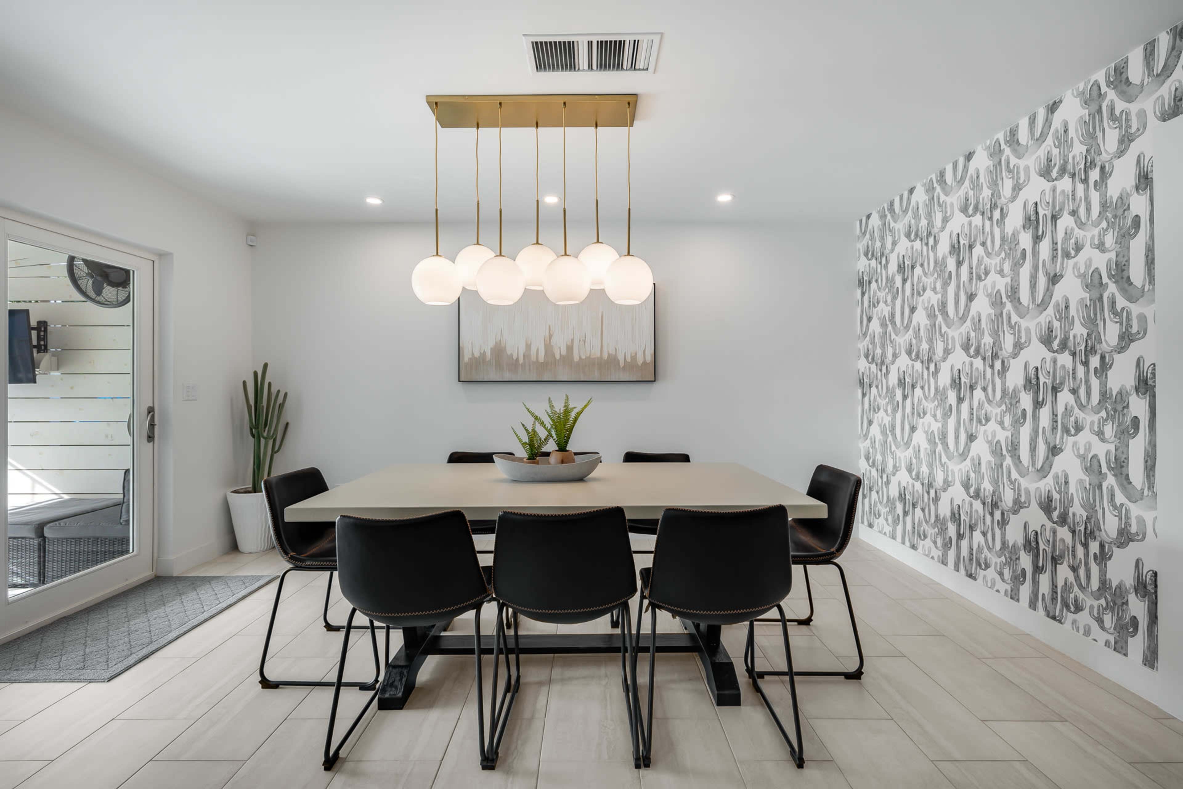A modern dining area features a large table surrounded by black chairs, with a decorative light fixture above and a cacti-patterned wallpaper on one wall.
