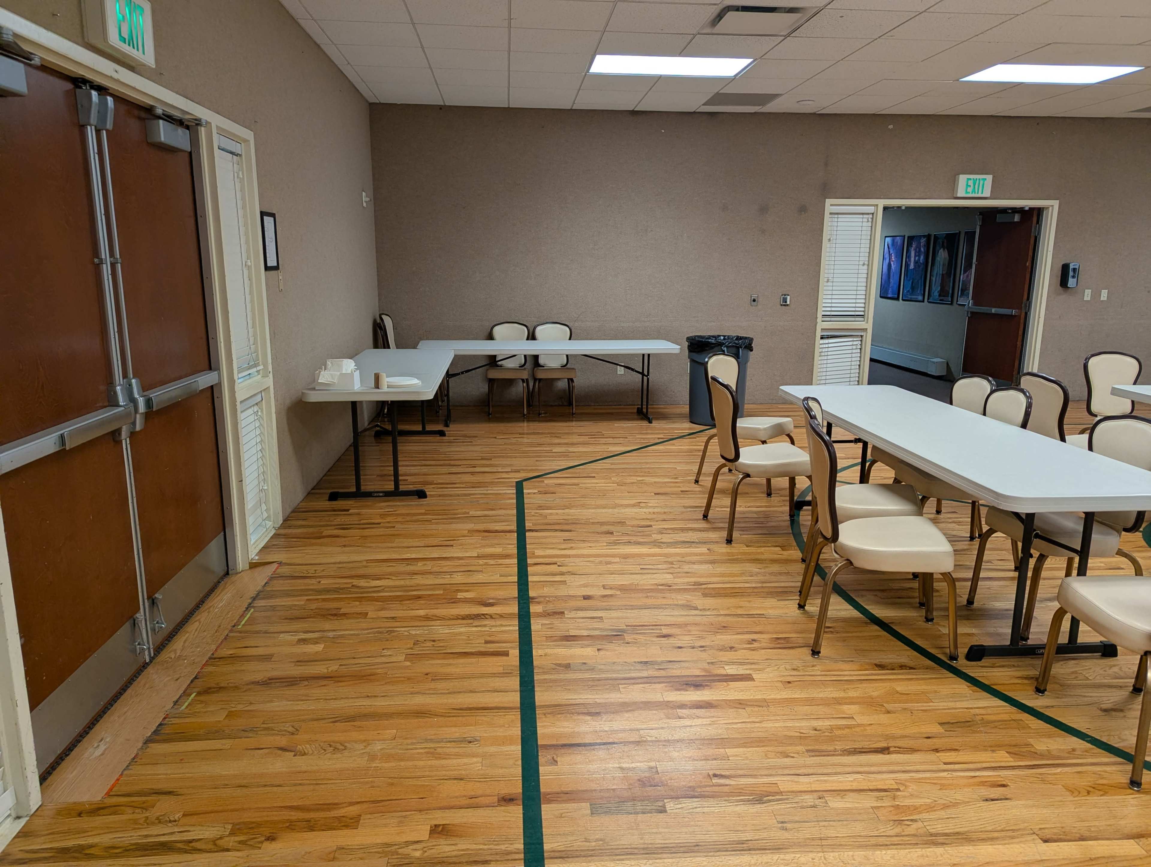 The image shows a large room with wooden floors, featuring a couple of tables and chairs arranged along the walls, with a trash can and a couple of doors visible.