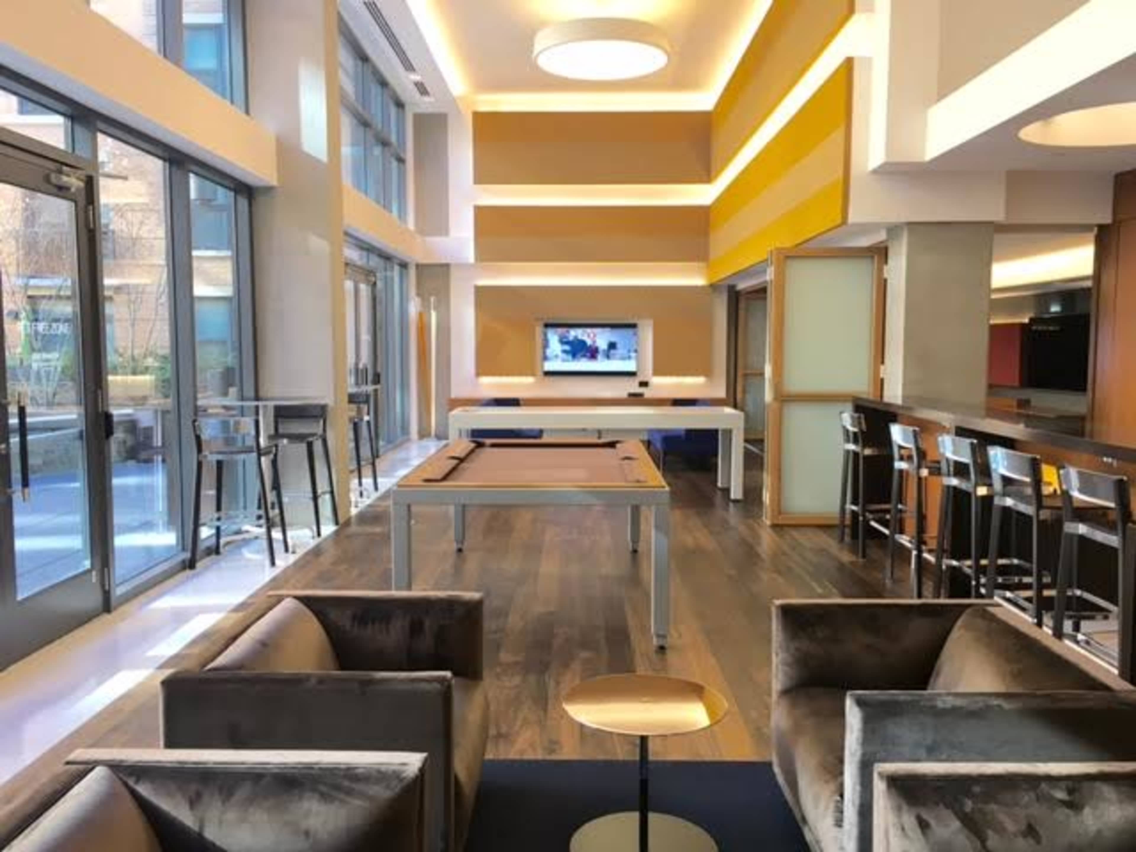 A modern lounge area featuring a pool table, seating arrangements, and a television mounted on the wall, with large windows allowing natural light to enter.