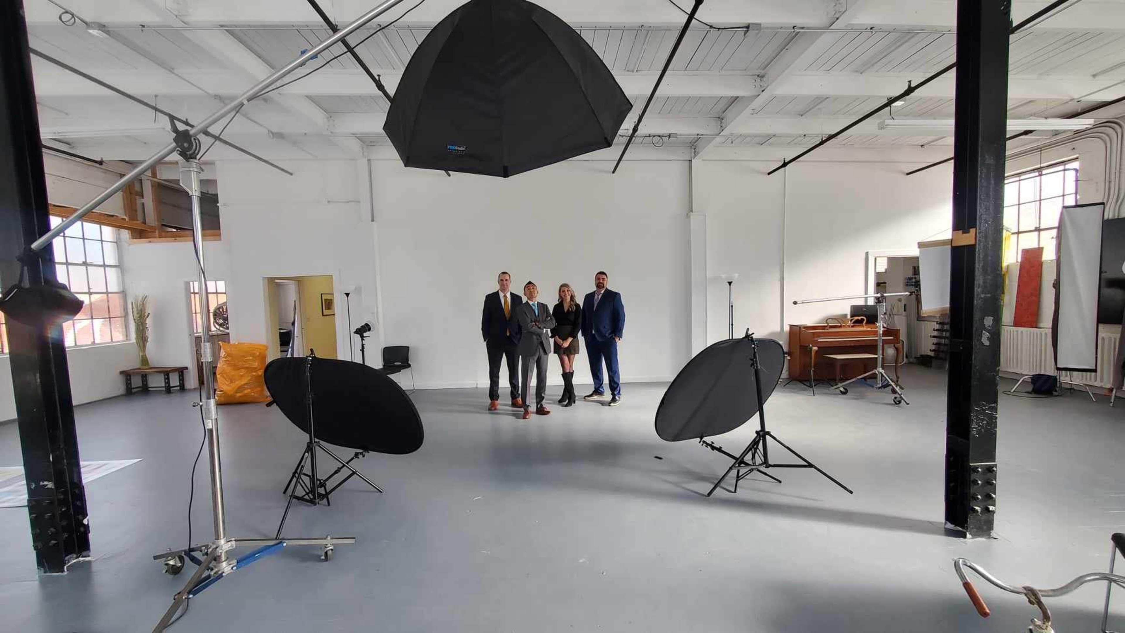 A group of three men in suits stands in a well-lit studio surrounded by photography equipment and backdrops.