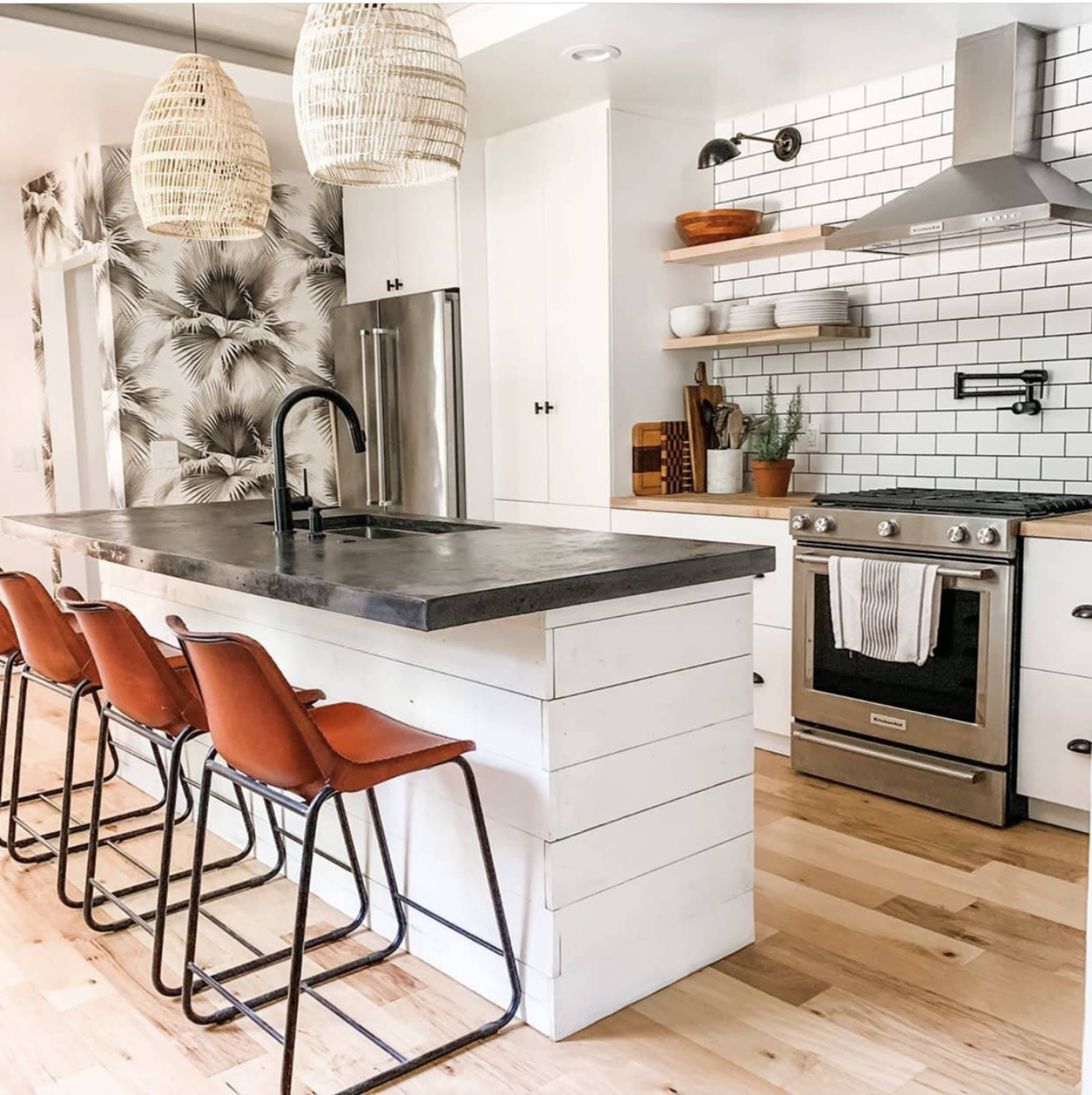 A modern kitchen features a large island with four metal-framed stools, a stainless steel oven, and a decorative wall of black and white tiles.