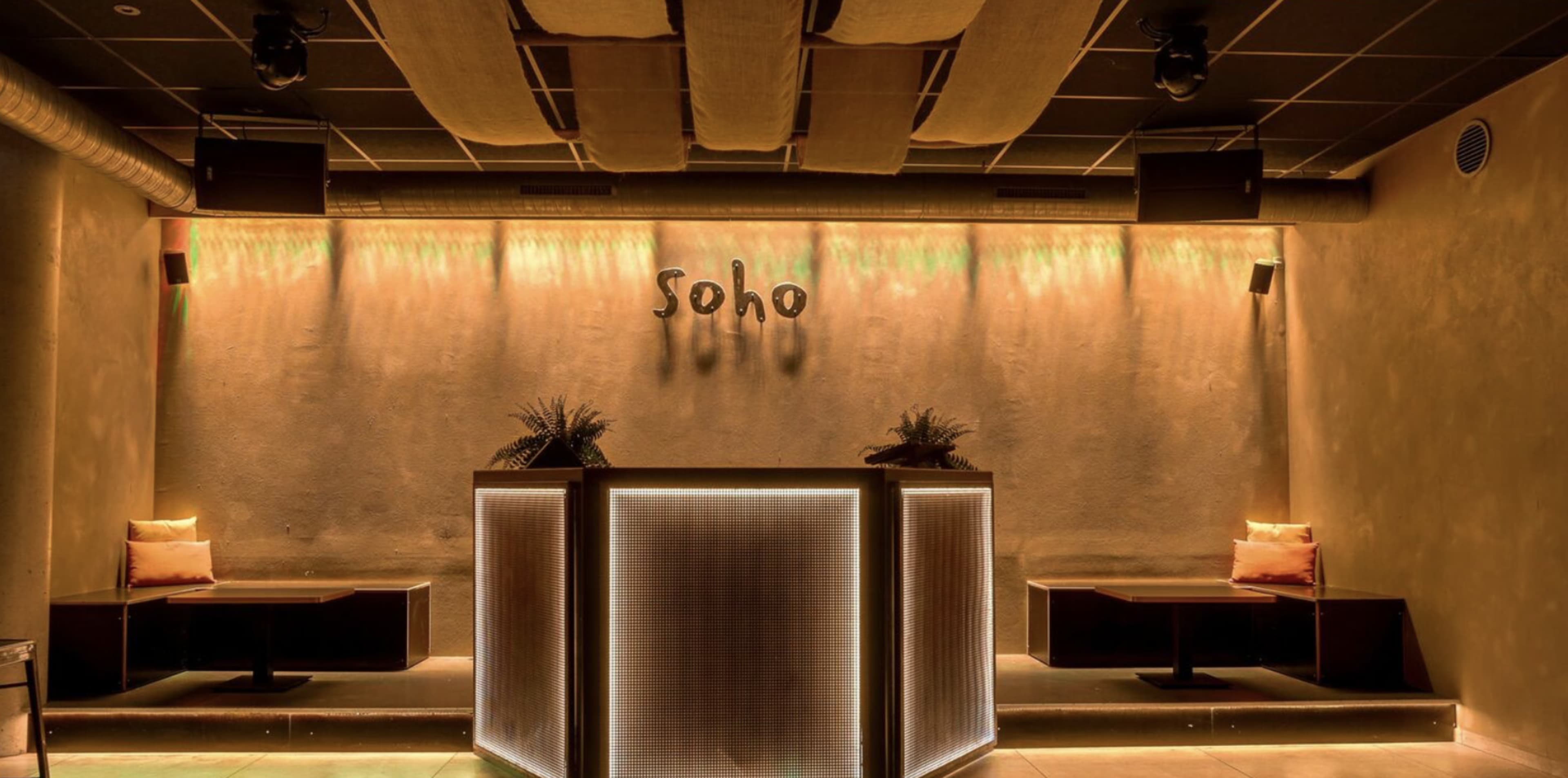 The image shows a dimly lit bar area with a modern design, featuring a counter illuminated by soft lighting and minimalist seating arrangements against a textured wall.