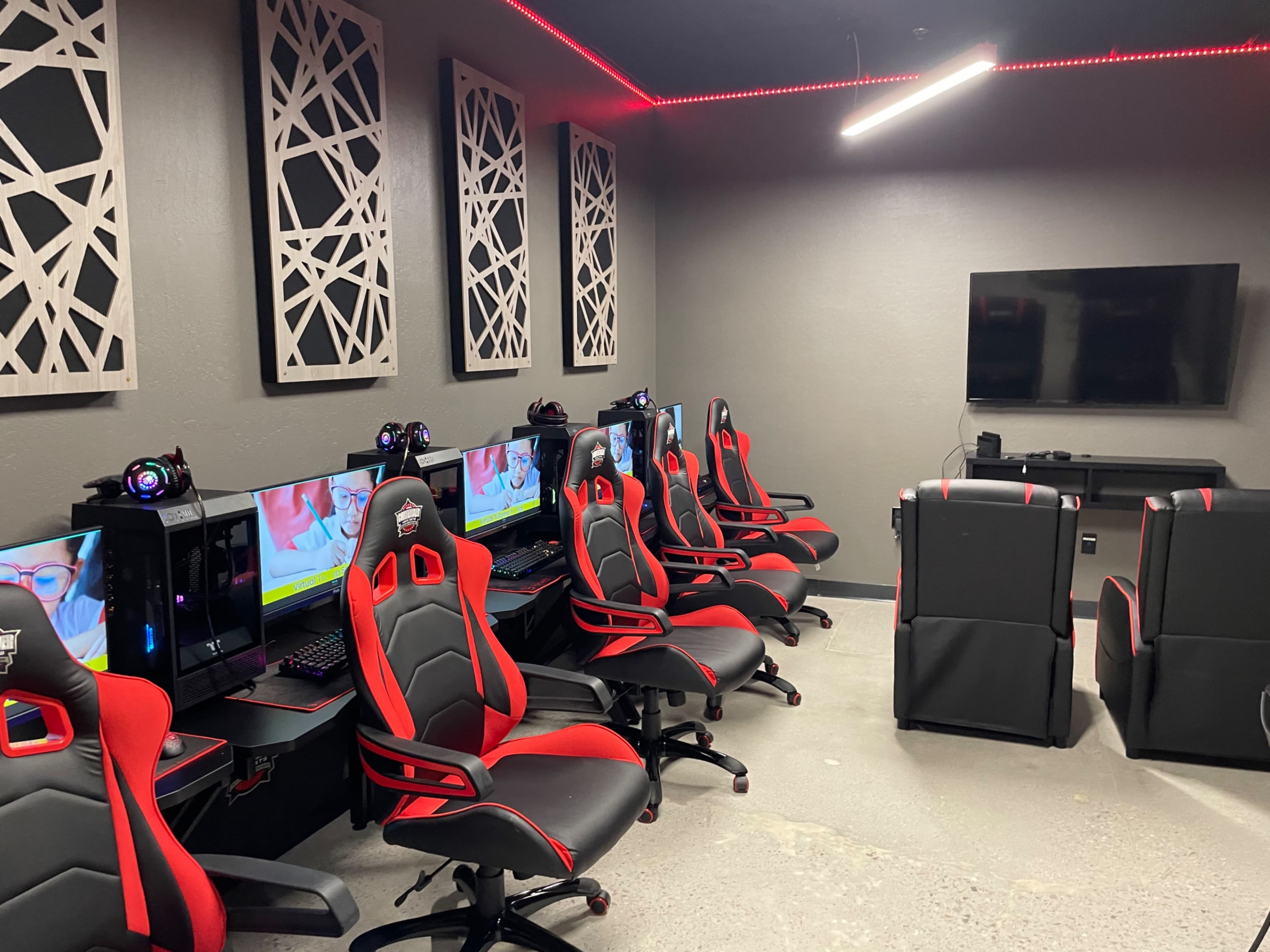 A gaming room equipped with multiple desktop computer setups and ergonomic chairs arranged along the walls.