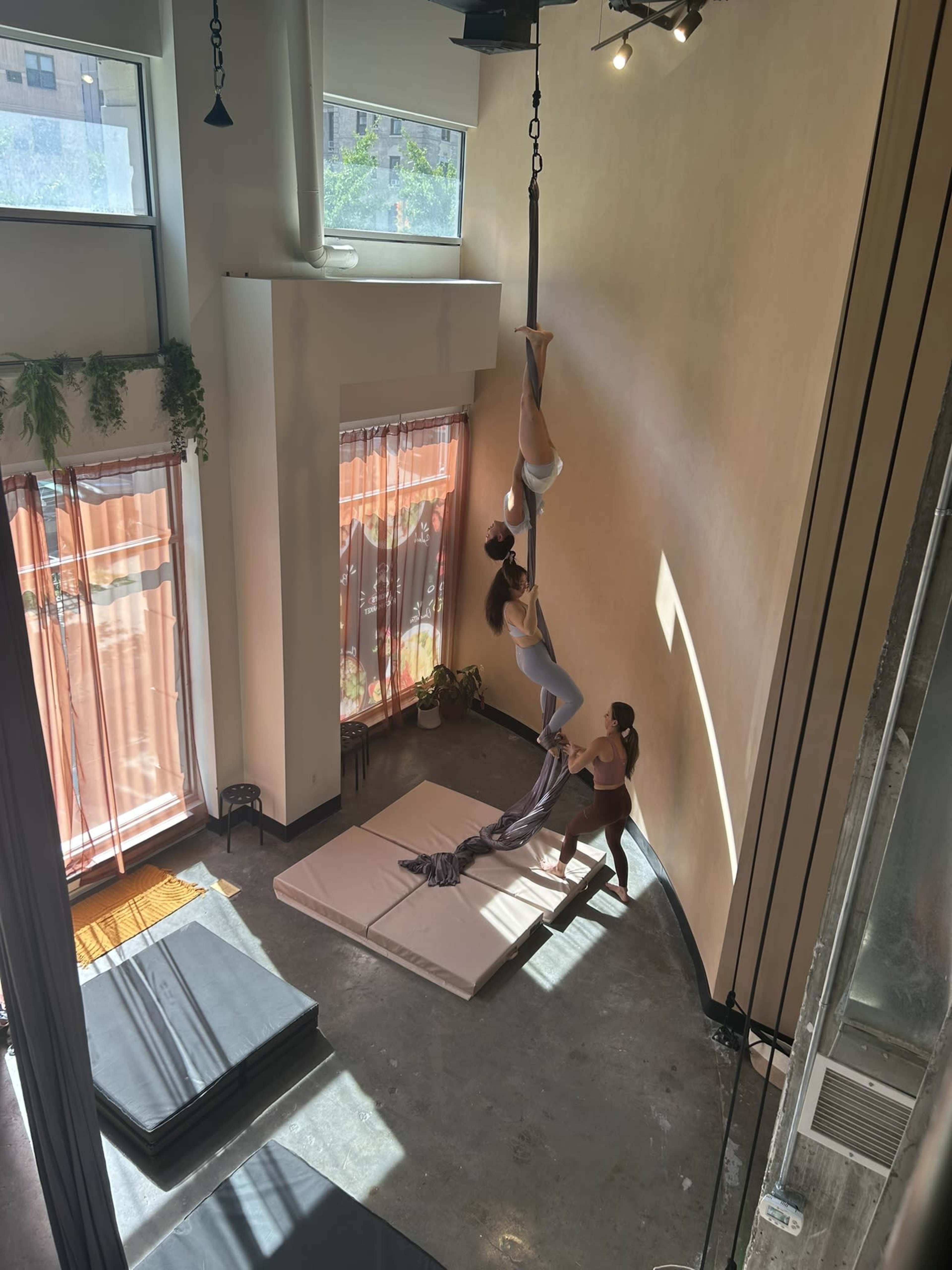 Two individuals practice aerial silks in a brightly lit studio with large windows and mats on the floor.