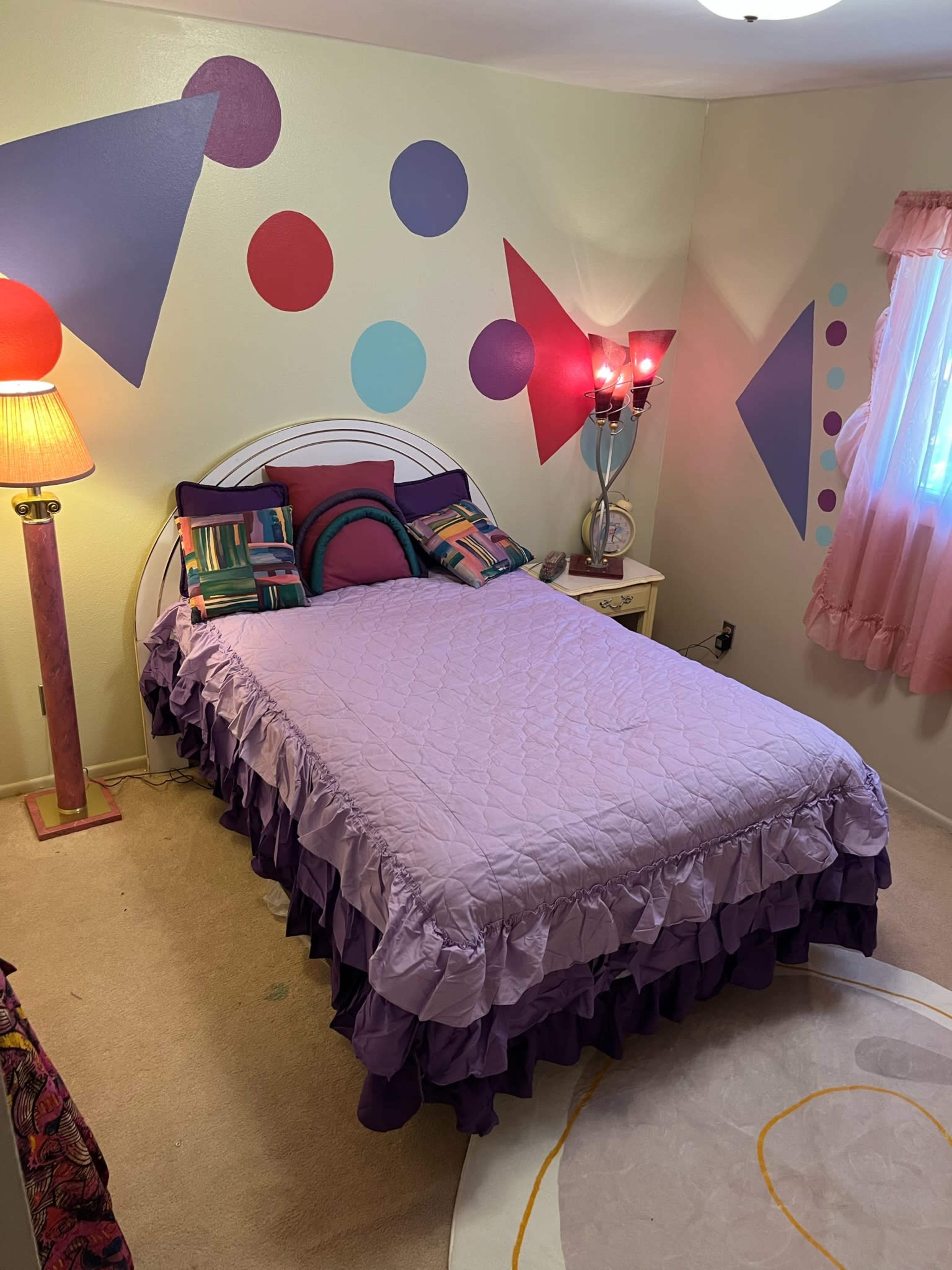 A bedroom with a lavender bedspread, decorative pillows, a lamp with a pink base, and colorful geometric wall art.